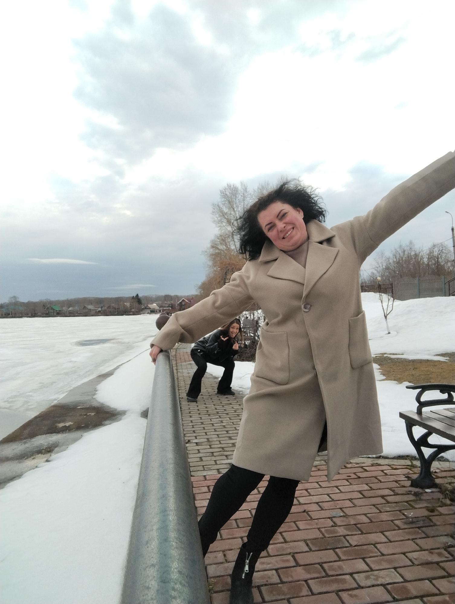 The image shows two women posing near a partially frozen body of water on a brick walkway.
The main subject is a woman in a tan coat, black leggings, and black boots, with her arms outstretched. She is leaning on a silver-colored metal railing. A second woman is crouched behind her on the walkway making a peace sign. In the background, there is a partially frozen river or lake with houses visible on the opposite bank. Snow is present on the ground. To the right is a park bench and some trees. The cloudy sky suggests it is a cool day.
Based on the information given, the location is most likely Votkinsk, Russia.