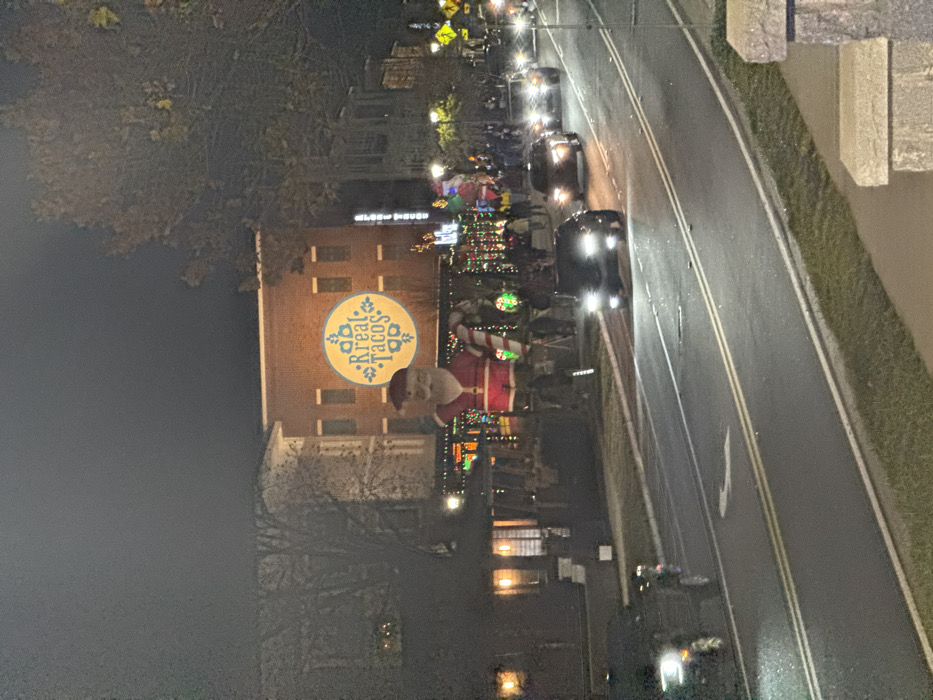 Night shot of a town street lined with Christmas lights. A building displays a circular "Real Tacos" sign and a large Santa Claus decoration. Cars drive on the right side of the road. Trees are partially visible on the left. The sky is dark, indicating nighttime. The overall scene conveys a festive holiday atmosphere.
