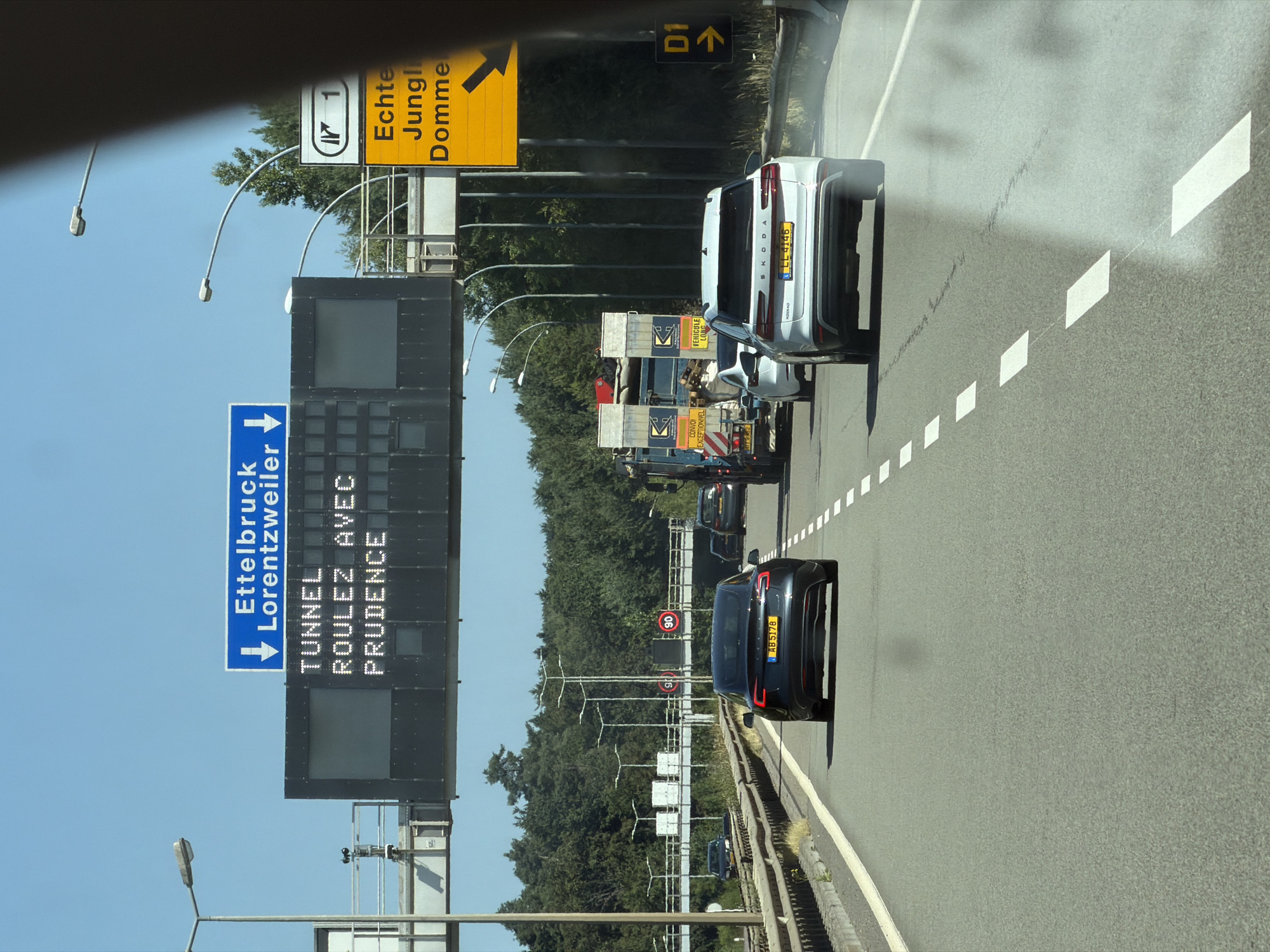 The image depicts a view from inside a vehicle traveling on a highway in Niederanven, Luxembourg. Overhead electronic signs display traffic information. The first sign, in blue, reads "Ettelbruck Lorentzweiler" with a downward arrow. Below this, a larger digital display shows "TUNNEL" in the top line and "ROULEZ AVEC PRUDENCE" in the second line, indicating a tunnel ahead and advising drivers to proceed with caution. To the right, a yellow directional sign indicates exit 1 and the destinations "Echternach Junglinster Dommeldange" with a rightward arrow. Various vehicles are visible on the multi-lane highway, including a black Tesla, a white Skoda, and several trucks. The environment is characterized by a clear blue sky, trees lining the highway, and streetlights. Speed limit signs of 90 km/h are posted on overhead gantries. The road surface is asphalt with clear lane markings.