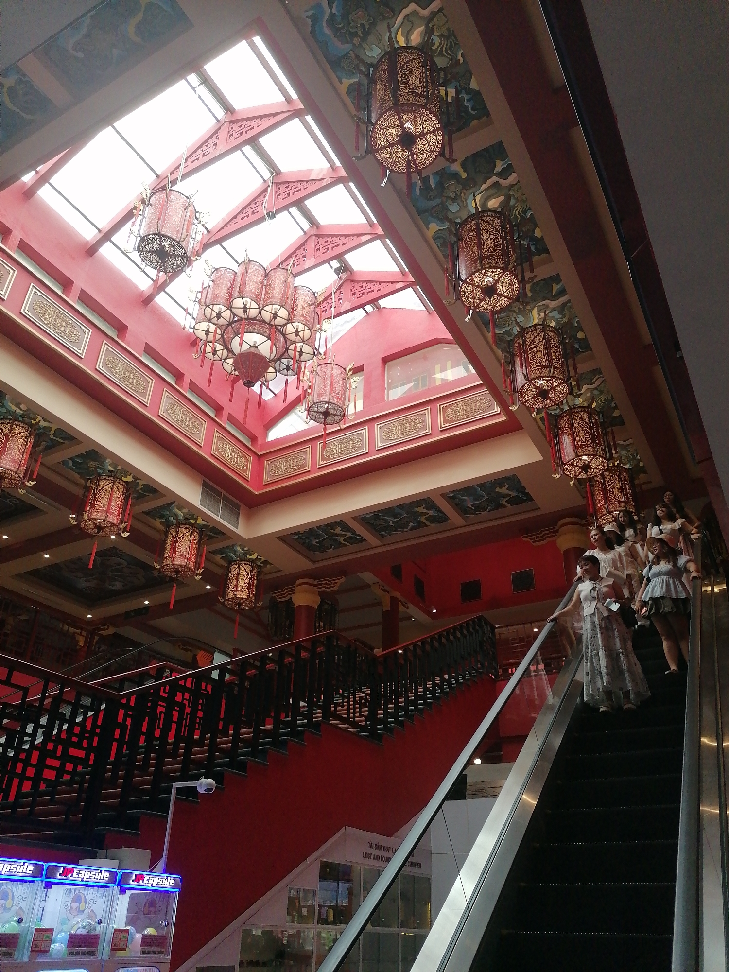 The scene depicts the interior of a multi-level shopping center in Bai Chay Ward, Vietnam. The dominant color scheme is red, accented with gold trim and black iron railings. Several red Chinese-style lanterns hang from the ceiling, which features a skylight. An escalator is visible on the right side of the image, with several women riding upwards. A staircase with black railings is located to the left. A row of capsule vending machines is visible at the bottom left. The overall architectural style suggests a theme park or cultural center.