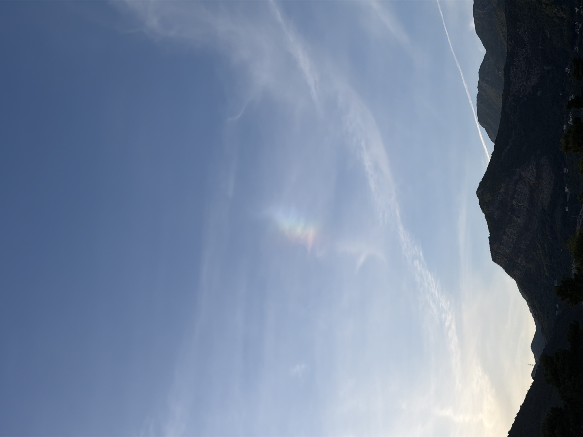 The image shows a view of the sky and mountains. The sky is predominantly blue with some wispy clouds. A faint rainbow-like iridescence is visible in the lower-center of the sky. On the right side, there are dark, rocky mountains with some trees. The location appears to be Budva, Montenegro. A faint white trail, possibly a contrail, is visible above the mountains.