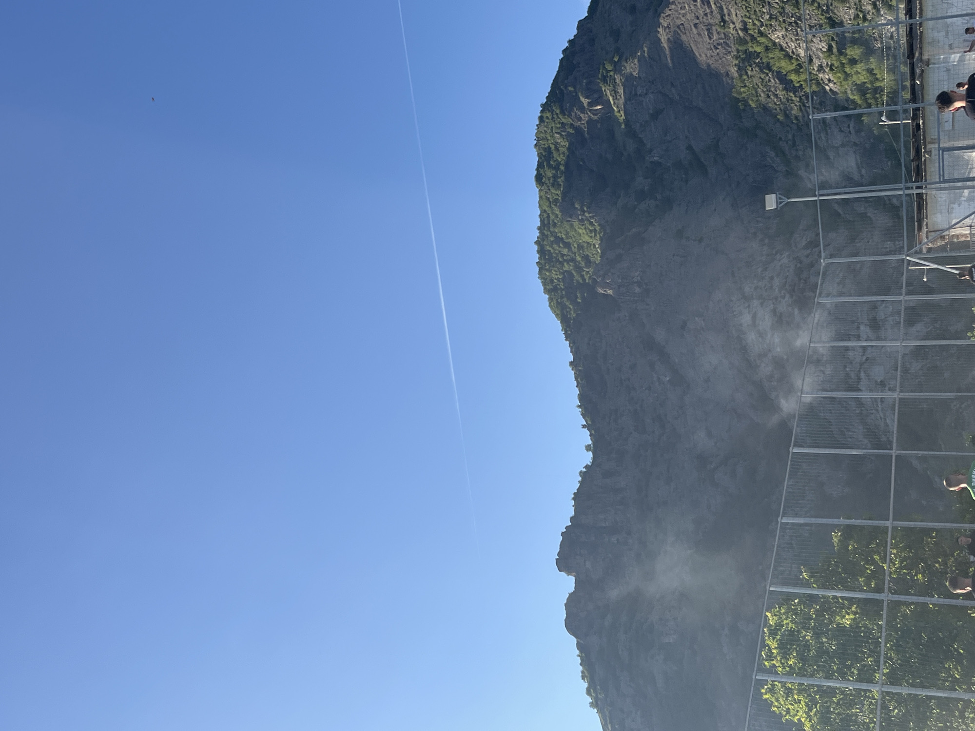 The image depicts a tall, rocky mountain in Bukovo, Bulgaria, partially covered with greenery and facing a clear, blue sky with a contrail. A metal structure resembling a fence or viewing platform is visible in the foreground, with glimpses of people standing on it. Mist or spray partially obscures the base of the mountain.