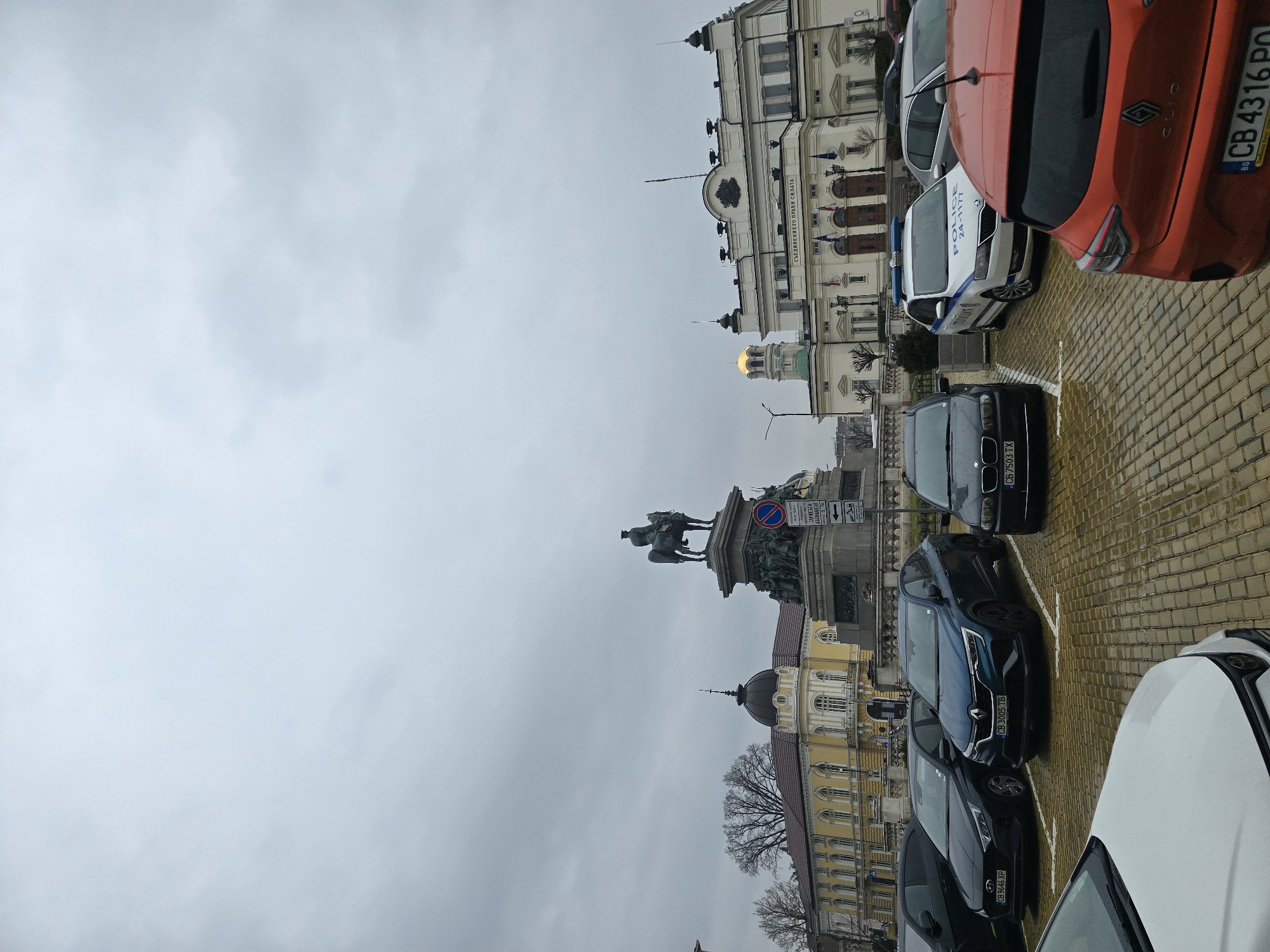 The image depicts a cityscape in Sofia, Bulgaria, likely a central square or public area. In the middle of the frame is the equestrian statue of Tsar Alexander II. The Bulgarian Parliament building and other governmental buildings are visible in the background. The sky is overcast. Cars are parked in the foreground. The ground is paved with paving stones, and the wet surface indicates it may have recently rained.