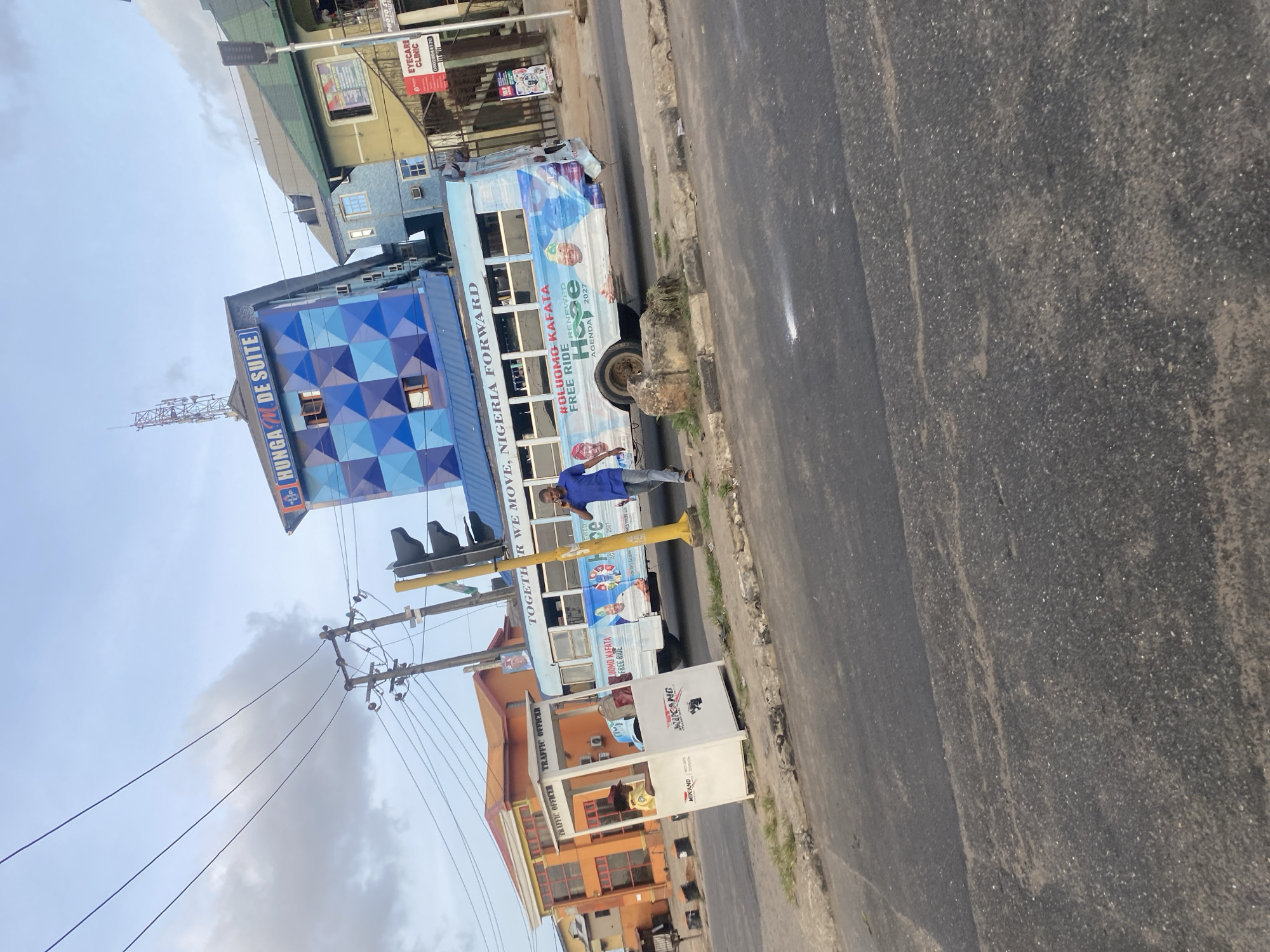 The image depicts a street scene in Agege, Nigeria. A bus advertising "Oluomo Kafata Free Ride, Renewed Agenda 2027" is parked near a traffic officer's booth labeled "Mikano". A man in a blue shirt is standing next to the bus. A building with "Hungam de Suite" signage is visible in the background, along with other buildings. Power lines and a traffic light structure are also present. The street is paved and shows signs of wear.