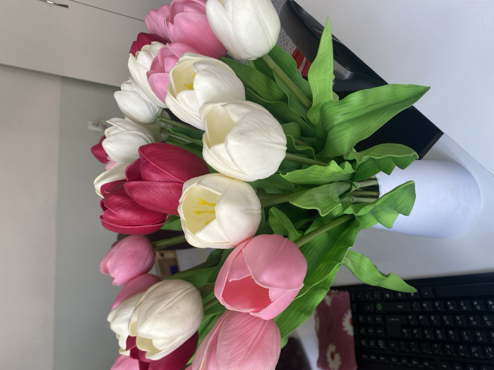 A vertical, overhead shot showcases a bouquet of artificial tulips in a white vase, set against a mirrored surface and a light-colored wall. The tulips are a mix of colors including white, pink, and a deep magenta. The arrangement is lush, with abundant green leaves.
The bouquet is positioned on a desk, with a black keyboard visible in the lower right corner of the frame. A hint of a dark rectangular object, possibly a laptop or monitor, is visible behind the flowers, suggesting an office or home workspace. The lighting appears to be artificial, casting a bright, even illumination on the scene, without any strong shadows, indicative of an indoor setting. The overall mood is serene and decorative, with the vibrant colors of the tulips providing a focal point. There is no discernible time of day or weather information, and the location cues are limited to a desk setup. No people are visible in the frame. The visible text from the keyboard is "N B M T K P m n W U H G H Y".