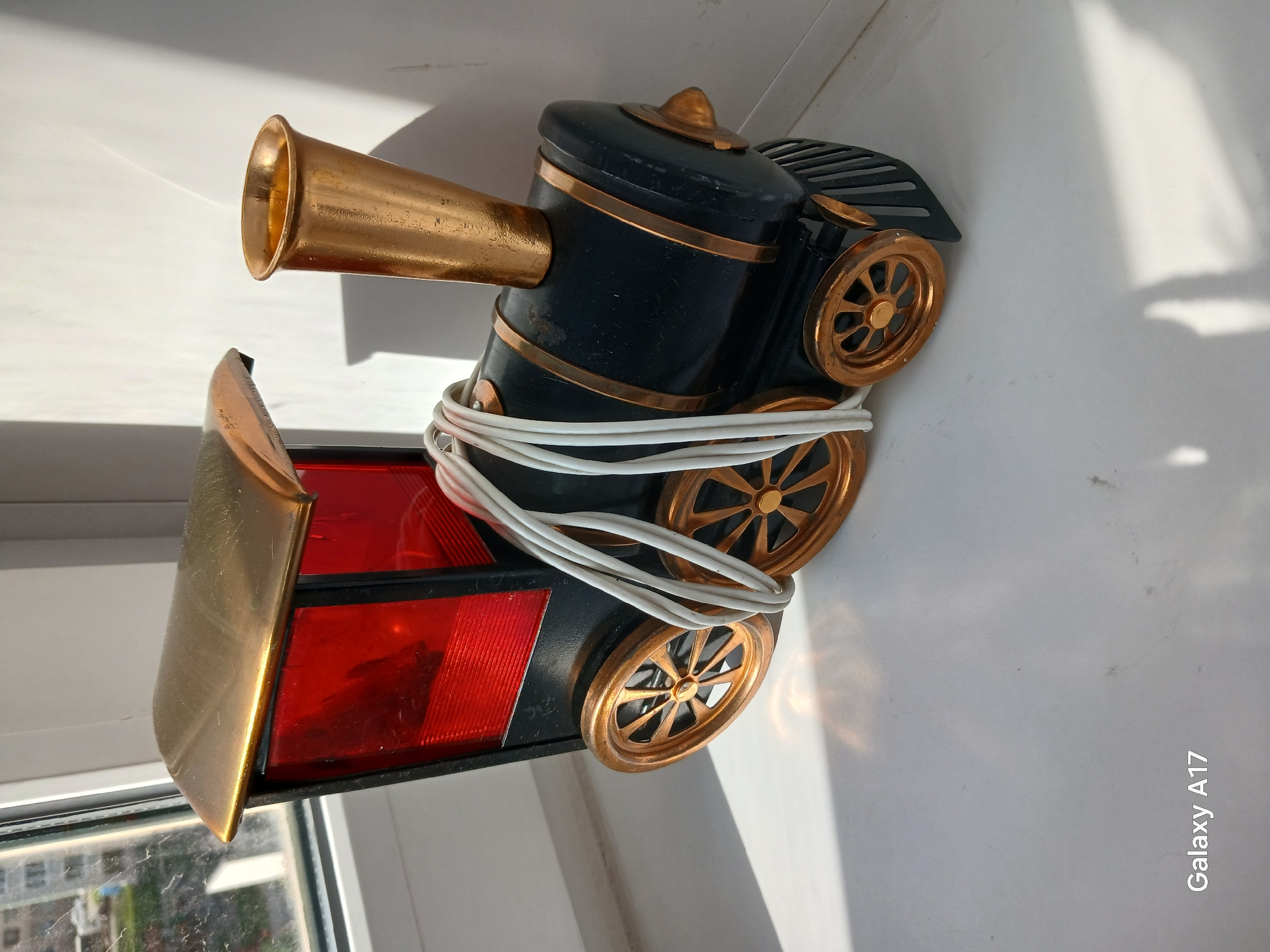 The image shows a toy train lamp with a black body and golden accents. The train has a large golden horn and a red and black caboose. A white electrical cord is wrapped around the body of the train. The train lamp is positioned on a white surface near a window. The window offers a glimpse of a cityscape, possibly in Yekaterinburg, Russia. The text "Galaxy A17" is visible in the bottom right corner of the image.