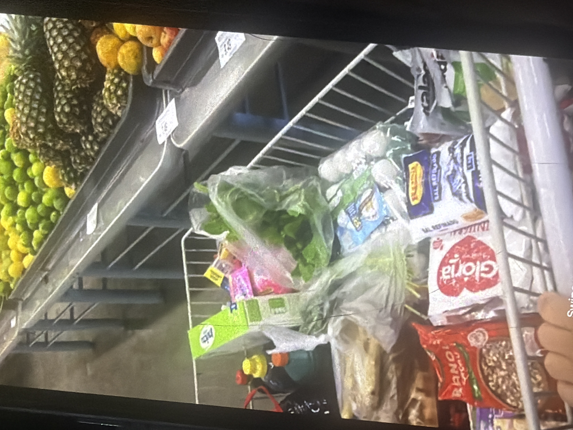This is a medium-shot, slightly tilted, interior photograph of a grocery store shelf stocked with various products. In the foreground, on the left side, there is a metal shopping cart with white bars. Inside the cart, there are several items: a red bag with the word "RANO" in white lettering, a white bag with a red heart and the word "Gloria" inside it, and a clear plastic bag containing white spherical objects. Behind these items, there are more bags and green leafy vegetables.
Moving to the right and slightly further back, the shelf extends horizontally. On this shelf, there are several items including a green box with white lettering that appears to be "PTS". Next to this, there are small colorful packages and more green vegetables. Further to the right, a tiered metal shelving unit is visible, with the top two shelves appearing empty and the lower shelves displaying products.
On the far right of the image, at the bottom, a display of fruits is present. There are pineapples with their spiky exteriors and leafy tops. To the right of the pineapples, there are piles of small, round, green fruits, possibly grapes or peas. In front of the pineapples and green fruits, there are a few round, yellow and orange fruits, possibly lemons or small apples. The overall environment suggests a well-stocked retail space, likely a supermarket or a local grocery store. The lighting appears to be artificial, typical of an indoor retail setting. There are no visible people in the frame. The image is focused on the products on display and the shelving units.