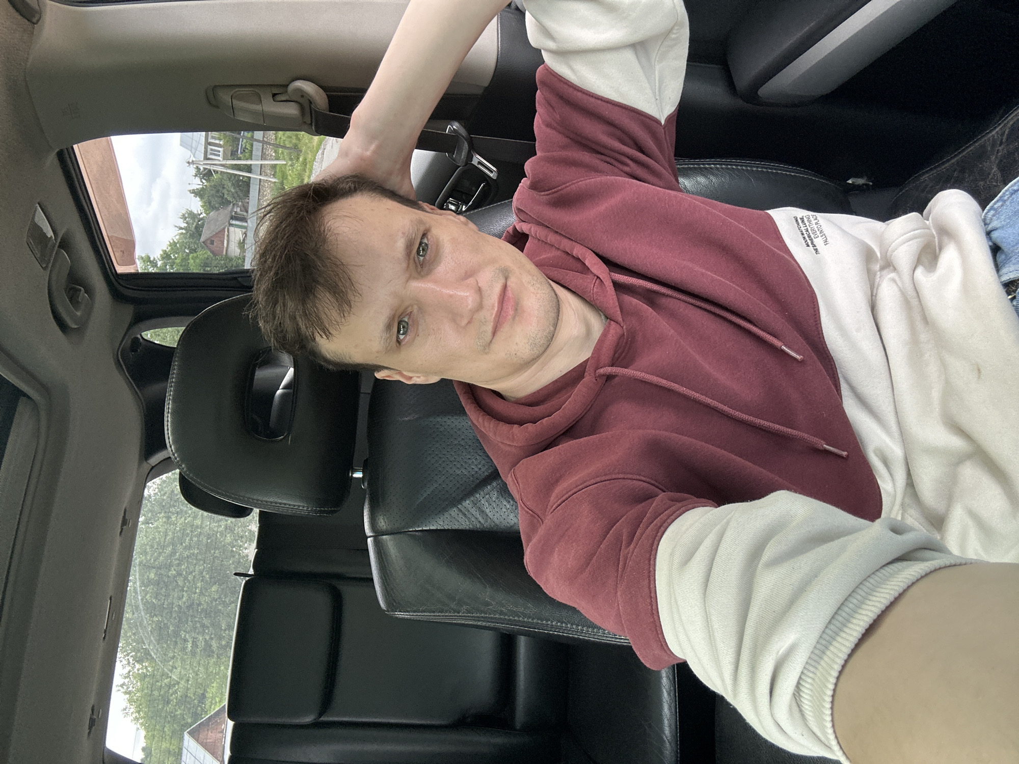 A young Caucasian man with light skin, light brown hair, and light green eyes is seated in the driver's seat of a car, taking a selfie. He is wearing a maroon and white hoodie with the sleeves rolled up. His right arm is bent at the elbow, with his hand resting on the back of his head. The car's interior is visible, including black leather seats and a seatbelt. Through the car window, a rural landscape can be seen with trees, houses, and a cloudy sky. The location is identified as Makiv, Ukraine.