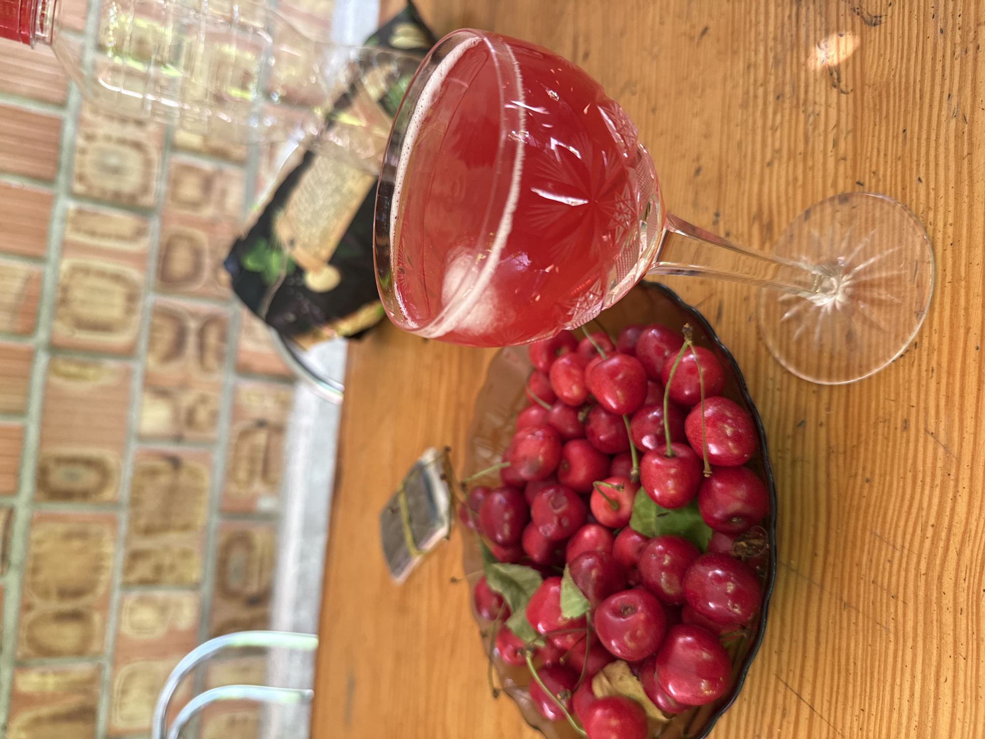 A high-angle, close-up shot displays a clear glass goblet filled with a vibrant red liquid, likely cherry juice or a similar beverage, adorned with a frothy white head. The goblet sits on a wooden table, its stem casting a shadow. Beside the goblet, a dark bowl overflows with fresh, ripe cherries, many still attached to their stems and some with green leaves. A small, dark object, possibly a wrapper or tag, lies near the cherries. In the background, a clear plastic bottle is partially visible, its cap a bright red. Behind the bottle, a section of a wall made of textured, beige tiles is visible, with faint shadows suggesting an outdoor setting. The wooden table's grain is prominent, indicating a rustic or natural environment. The lighting suggests it is daytime, possibly outdoors given the tiled wall and the general feel of the scene. The overall impression is one of refreshment and abundance, highlighting the fresh fruit and the inviting drink.