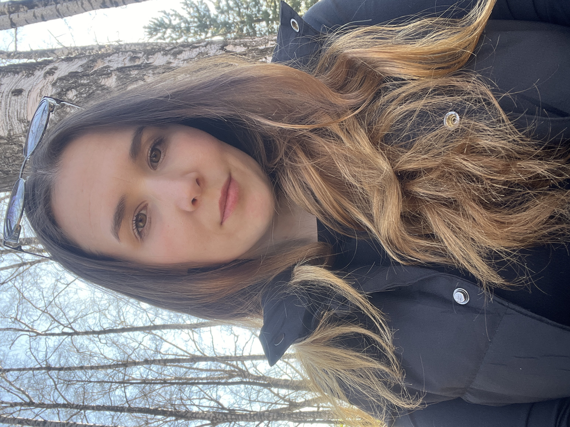 A selfie of a woman in front of a birch tree with bare branches. She is wearing a black puffy jacket and has sunglasses perched on her head. The city of Votkinsk, Russia, is not discernible from the image.