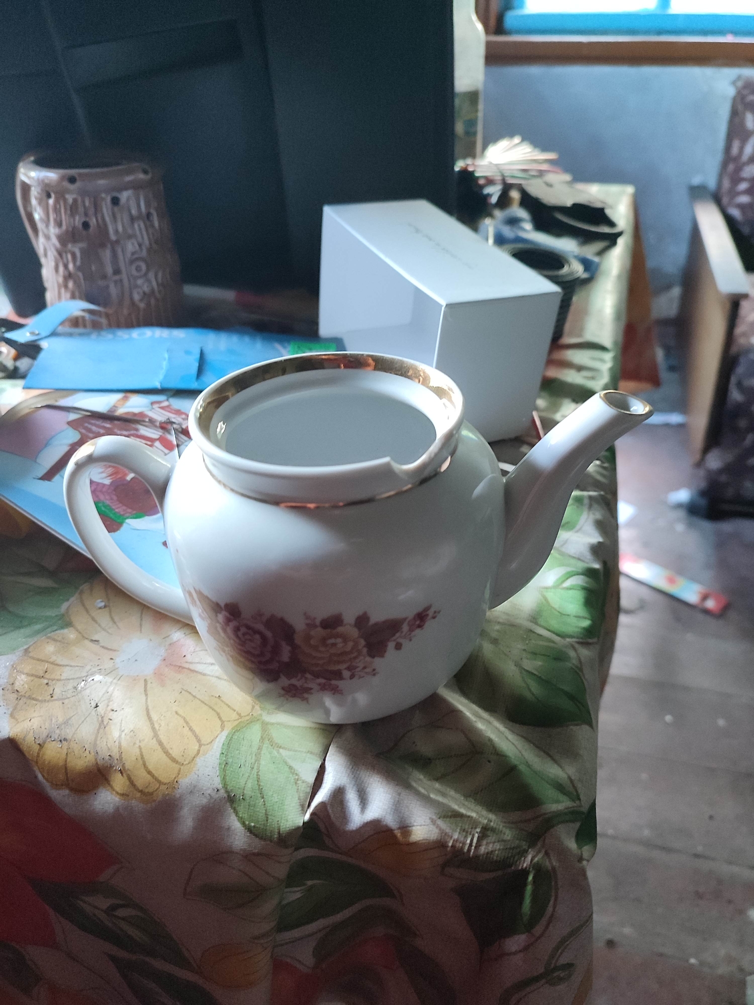 An indoor, high-angle, full shot captures a white ceramic teapot with a gold rim and floral pattern. The teapot sits on a floral-patterned tablecloth, which is draped over a surface. To the left of the teapot, a blue package is visible. In the background, a brown ceramic mug with an embossed pattern is partially visible. Behind the mug and to the right, a white rectangular box and a stack of papers are present. Further in the background, a wooden chair and a window with a blue frame are discernible. The scene appears to be set within a room in Gremyachinsk, Russia.