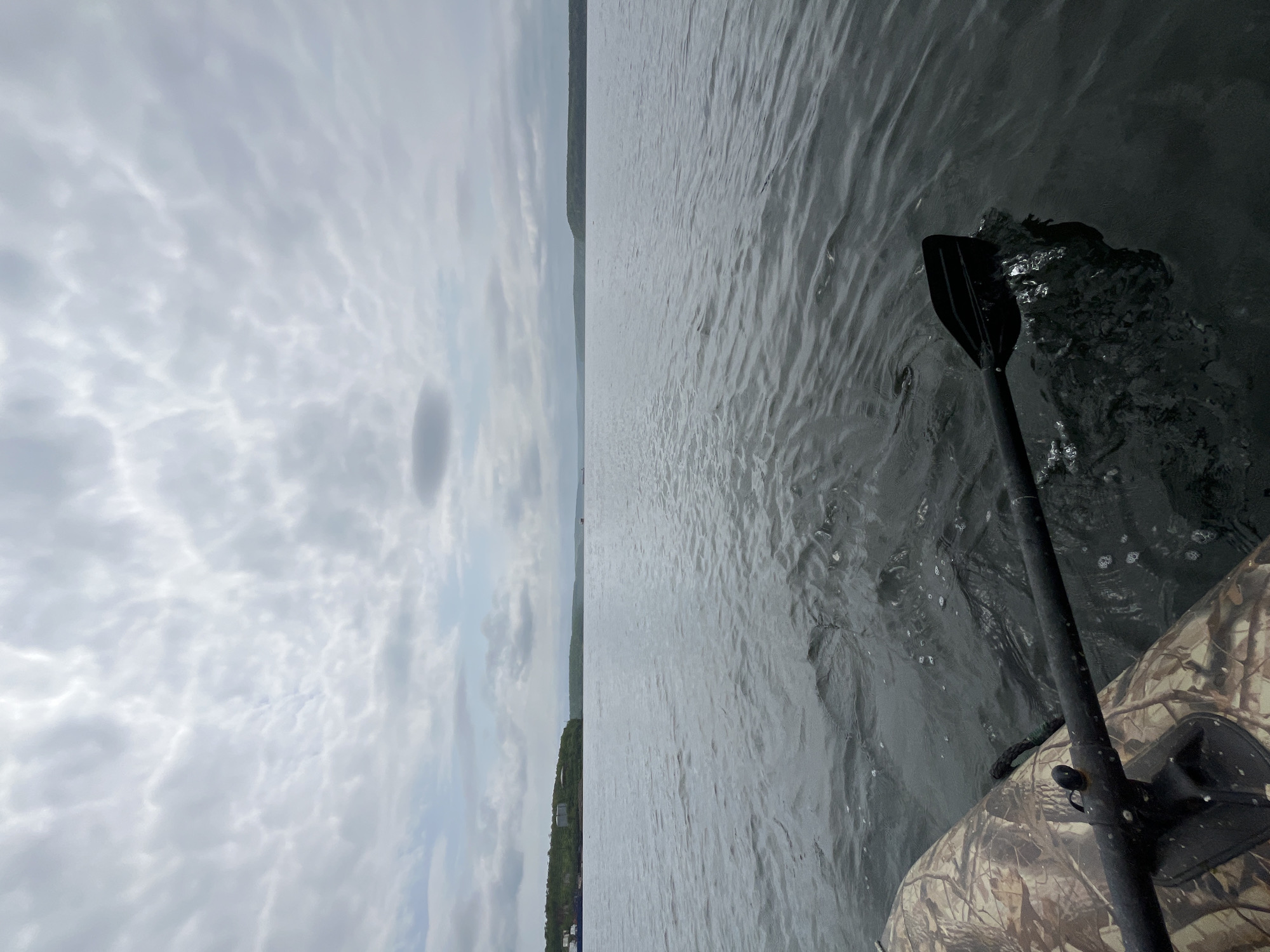 The image shows a first-person view from a camouflage-patterned inflatable boat on a calm body of water, likely a bay or inlet. A black oar is partially submerged in the water on the right side of the frame, creating ripples and small splashes. The water appears dark and slightly choppy. In the distance, a tree-covered shoreline with some buildings is visible on the left. The sky is overcast with dramatic, textured clouds. The scene is set in Sovetskaya Gavan', Russia, a port city known for its maritime activities.