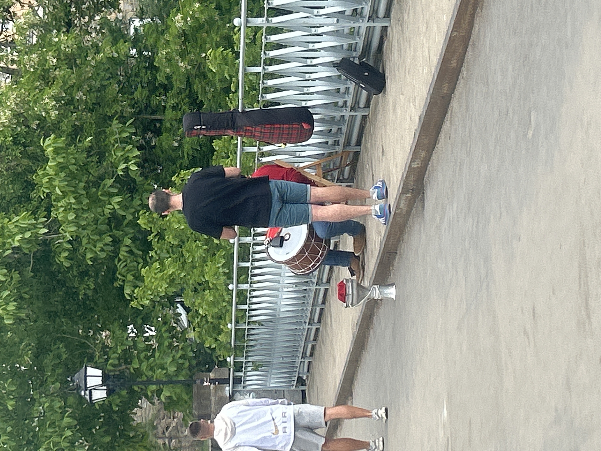 The image depicts a scene on a bridge in Veliko Tarnovo, Bulgaria. Two men stand on the pedestrian path. One man wears a black t-shirt, blue shorts, and blue sneakers. He stands near a drum and a red and black plaid guitar case. A second man wears a white sweatshirt with "AIR" printed on it, gray shorts, and white sneakers, standing at the bottom of the frame. The metal bridge railing is visible behind the man in the black t-shirt. Trees fill the background on the left side of the image. A black bag also sits on the bridge behind the man in the black t-shirt. A street lamp stands behind the second man.