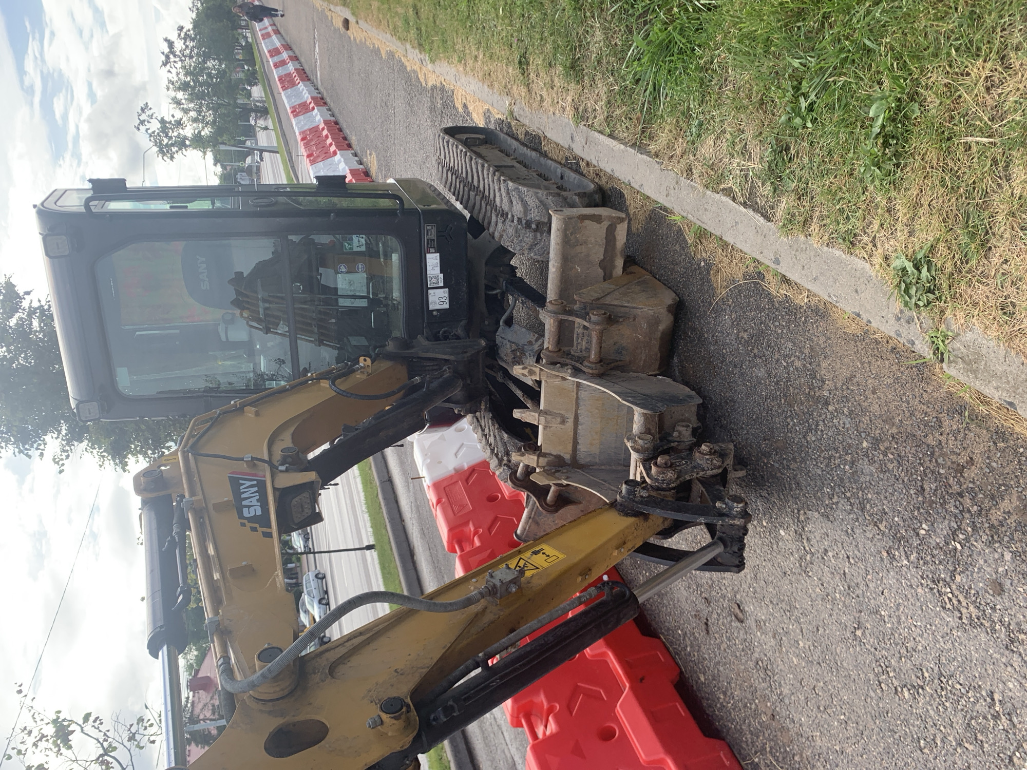 An excavator is parked on a paved surface, likely a road or construction site, next to a grassy area with a concrete curb. Red and white plastic barriers are placed in a line behind the excavator, separating it from what appears to be a street with cars and trees in the background. The sky is cloudy. The excavator is yellow and black, with "SANY" branding visible on its arm. There are no people immediately near the excavator, but a lone pedestrian can be seen walking on the sidewalk in the distance, past the barriers. The scene suggests ongoing construction or roadwork in an urban environment. Based on visual cues, this image is likely from Minsk, Belarus.
