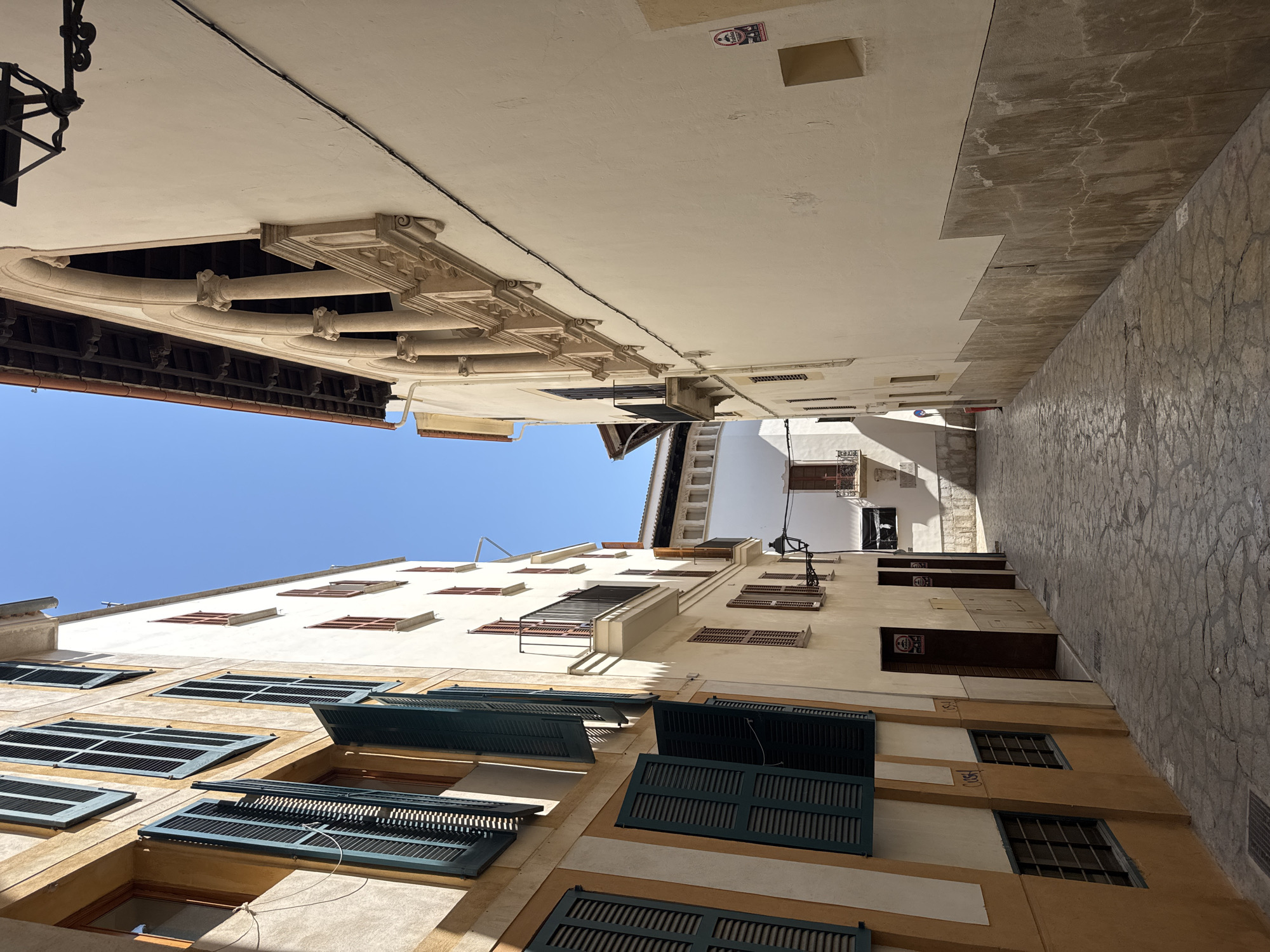The image shows a narrow, cobblestone street in the city of Palma de Mallorca, Spain, under a clear blue sky. Tall buildings flank both sides of the street, casting long shadows. The building on the left has a yellow and white facade with multiple windows covered by dark green shutters, some of which are open. The building on the right is cream-colored with a stone balustrade along an upper-level balcony supported by arched columns. A black wrought-iron street lamp is attached to this building. Several doors are visible on the ground floor of the left building, and a red trash bin is visible further down the street on the right. The architecture suggests a historic European city.