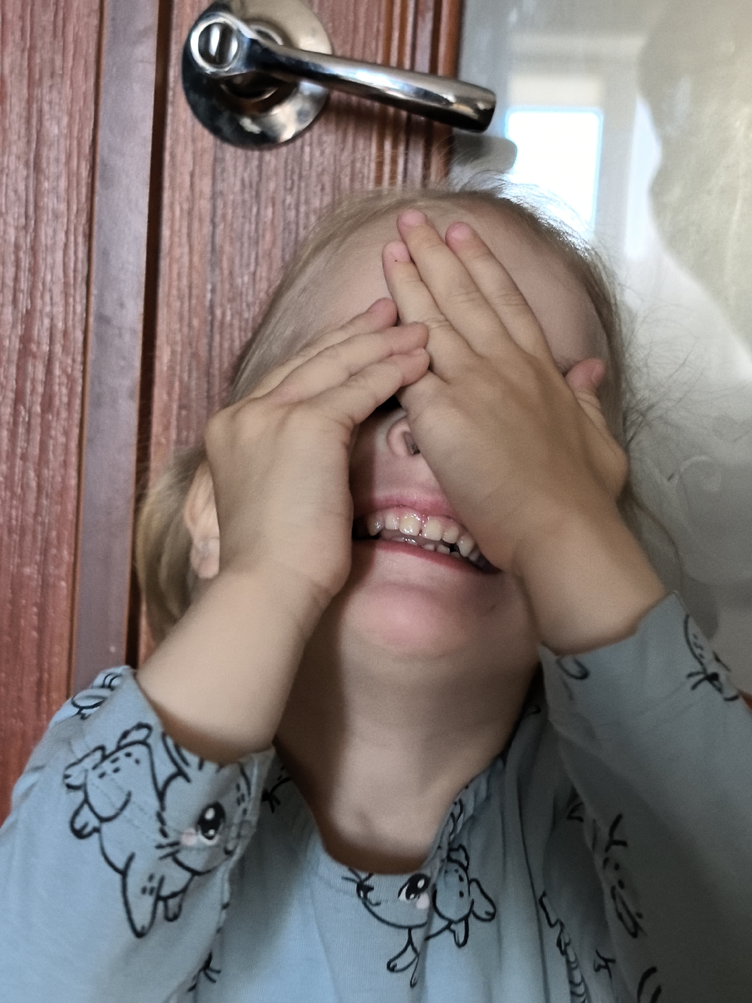 A young child with blonde hair is covering their eyes with both hands and smiling. They are wearing a light blue pajama top with a pattern of cartoon animals. The child is positioned in front of a wooden door with a silver door handle. The background is softly lit and out of focus, suggesting an indoor environment.