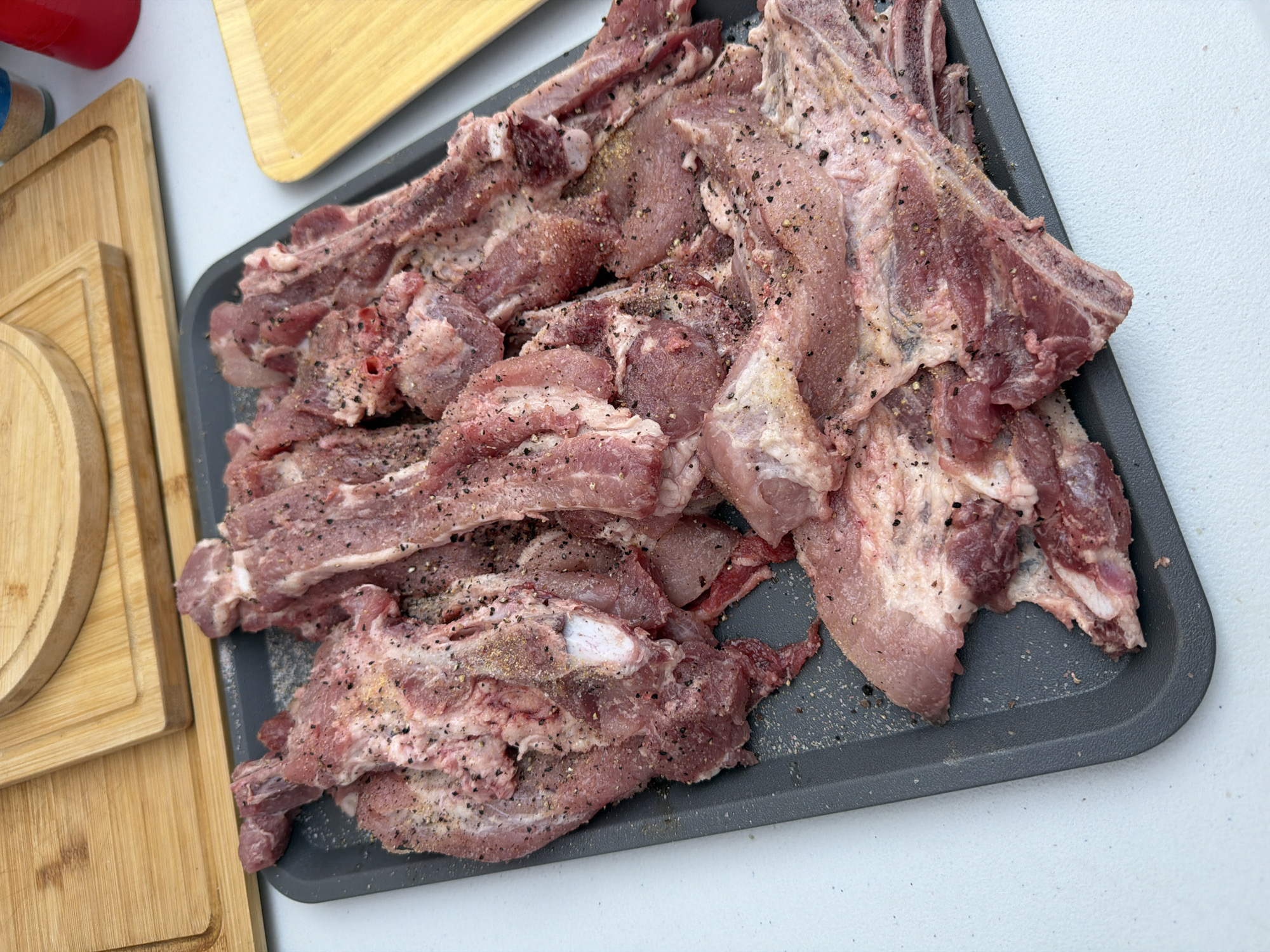 The image shows a grey tray filled with raw, seasoned pork ribs. The ribs are arranged haphazardly on the tray and are sprinkled with what appears to be black pepper and possibly other seasonings. The tray sits on a white surface, possibly a table. To the left of the tray, there are multiple wooden cutting boards stacked together. In the background, a red container is visible. The location is not discernible from the image. No people are visible.