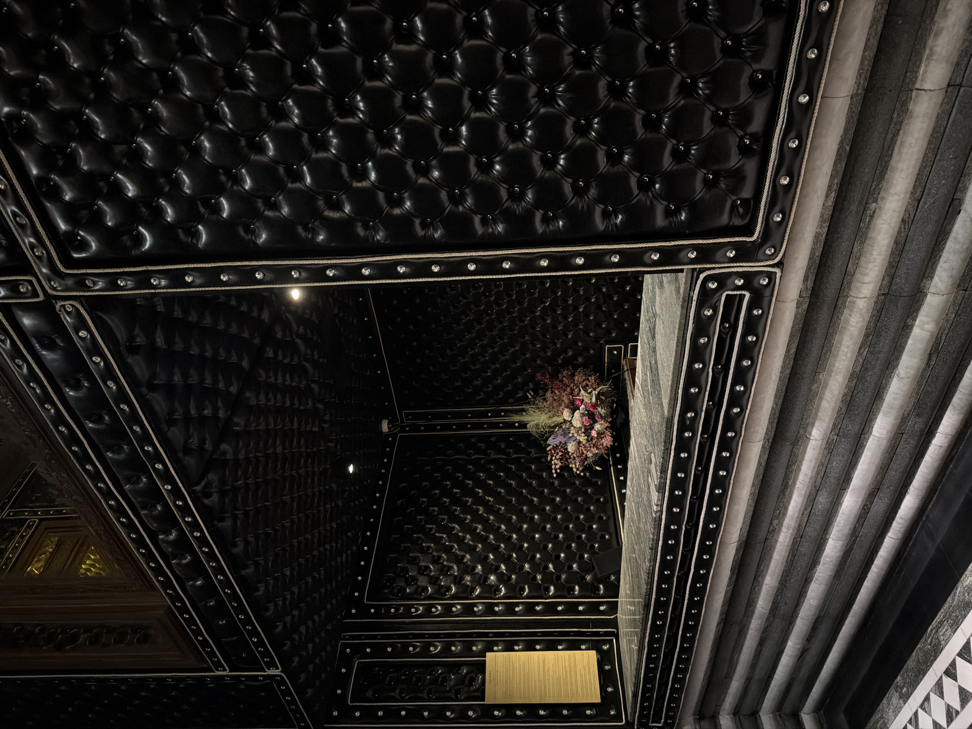 The image depicts the interior of a room in an unknown location. The walls and ceiling are covered in black tufted material, accented with white rope and jeweled studs. One wall is constructed of gray striped stone. A bouquet of flowers sits on the wall near the stone. A menu sits on the wall near the bottom of the frame.