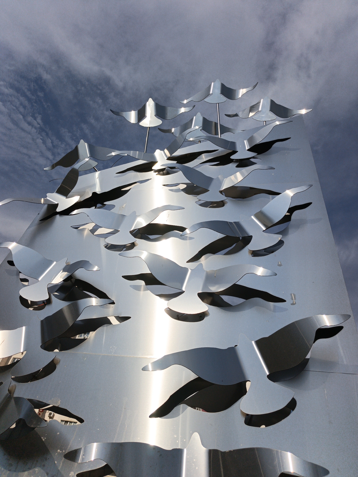 This media file features a large, metallic sculpture of birds in flight, mounted on a flat, reflective surface that appears to be part of a building or monument. The birds are abstractly rendered, with cut-out shapes that create shadows and depth. They are arranged in a dynamic composition, suggesting movement and a flock in motion. The sculpture is predominantly silver or brushed aluminum, reflecting the sky and surrounding environment.
The sky above is a mix of blue and gray clouds, indicating a partly cloudy day. The lighting suggests it is daytime, possibly late morning or early afternoon, given the bright reflections on the metal. The scene is outdoors, and the reflective nature of the sculpture indicates a polished surface, possibly stainless steel. There are no people visible in the image. The overall impression is one of modern public art, evoking a sense of freedom and natural beauty through stylized forms. The location context is Goryachiy Klyuch, Russia. No visible text is present in the image.