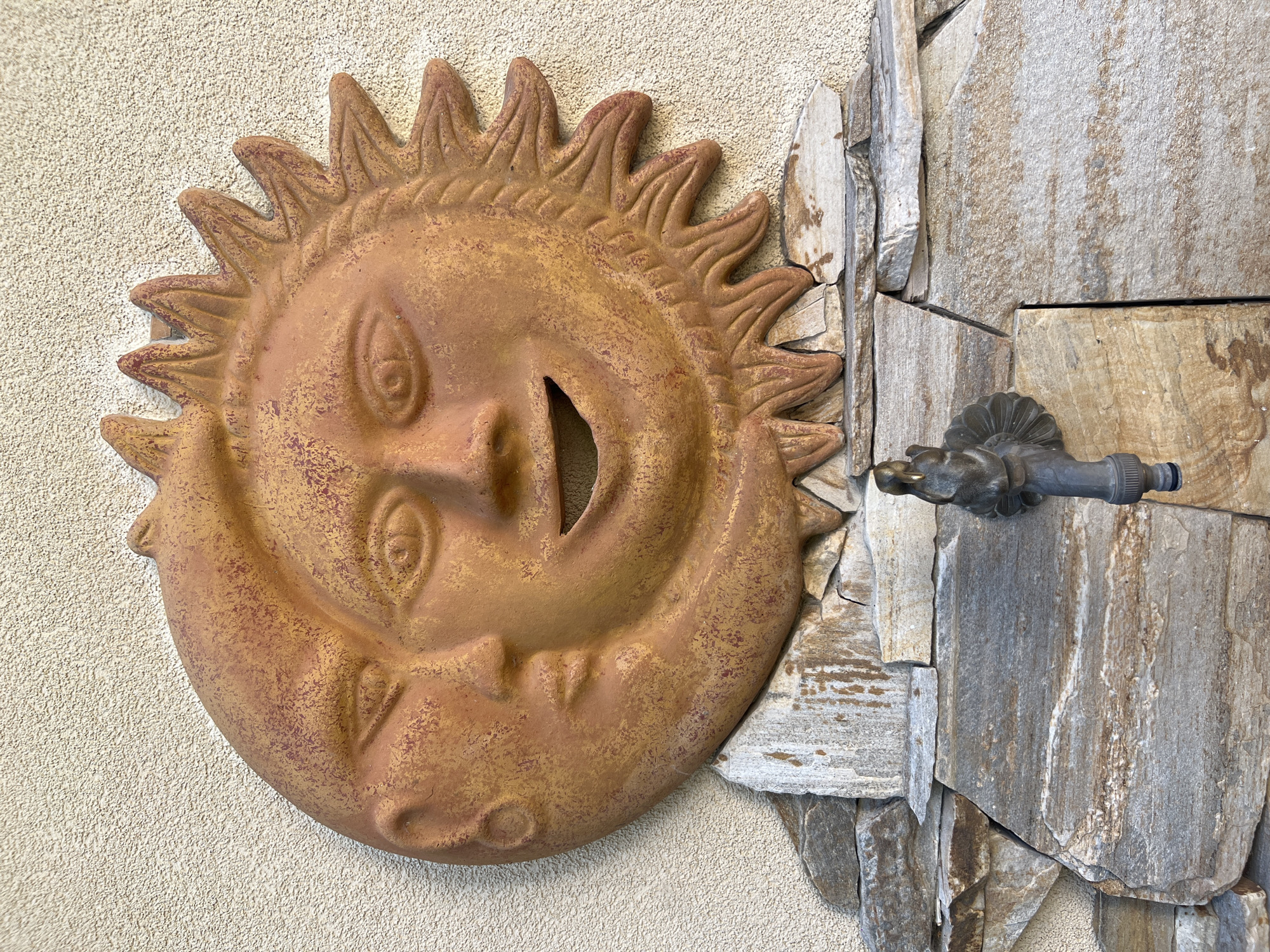 A decorative terracotta wall hanging depicting a sun and moon faces is mounted on a textured beige wall, situated above a stone facade. The sun face, on the right, is characterized by a wide open mouth and sharp rays. The moon face, on the left, has closed eyes and a serene expression. A brass water faucet with an ornate handle is attached to the stone facade below the wall hanging. The scene appears to be an outdoor setting, potentially a garden or courtyard.