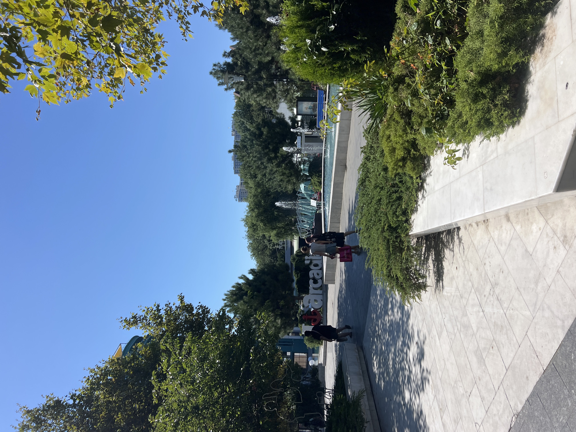 The image depicts a sunny day scene in Odesa, Ukraine, specifically within what appears to be a public plaza or park area with the large, white, illuminated sign "Arcadia" prominently displayed. Several people are visible walking on a paved pathway. In the background, there is a water fountain feature with jets of water spraying into a pool, surrounded by lush green trees and bushes. Buildings and structures, including some with multiple stories, are visible further in the distance, suggesting an urban setting. The foreground is dominated by paved surfaces and manicured greenery, indicative of a well-maintained public space.