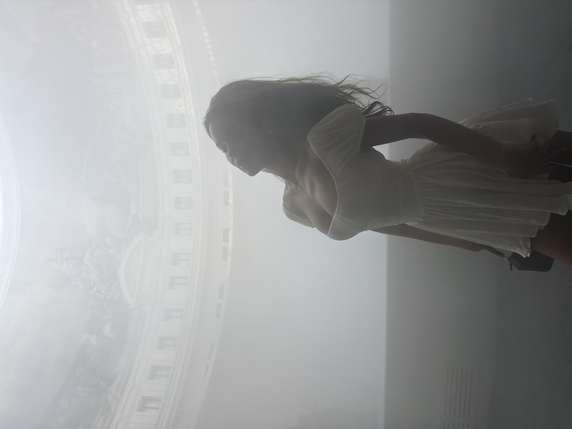 A woman in a white dress stands with her head tilted back, looking upwards. She holds a dark handbag. The background appears to be a large, domed interior space, possibly a museum or public building. The scene is filled with a bright, diffused light, creating a hazy, ethereal atmosphere. The architecture visible in the background suggests a classical style with curved walls and detailed ornamentation. The painting on the dome is obscured by the light and haze.