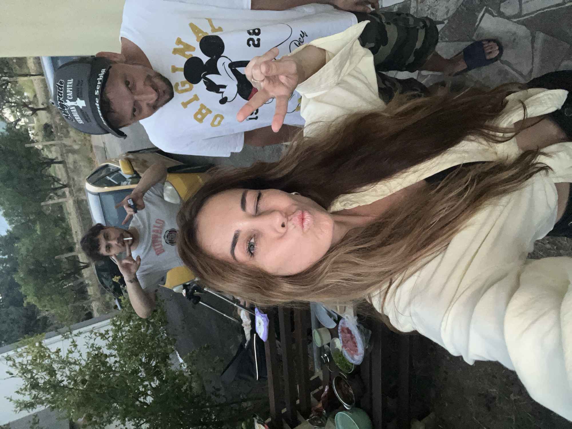 In the city of Bar, Montenegro, a close-up selfie shows three individuals. A woman in the foreground, with long brown hair and wearing a light-colored top, puckers her lips towards the camera, with her right eye winked. Behind her, a man in a grey t-shirt with "Buffalo" printed on it, holds two fingers up in a peace sign and has a cigarette in his mouth. To the right, another man wearing a black cap that reads "Belgrade Serbian Capital" and a white t-shirt with a Mickey Mouse graphic, smiles at the camera and makes a peace sign. They are outdoors, with a table laden with food, a car, and greenery visible in the background.