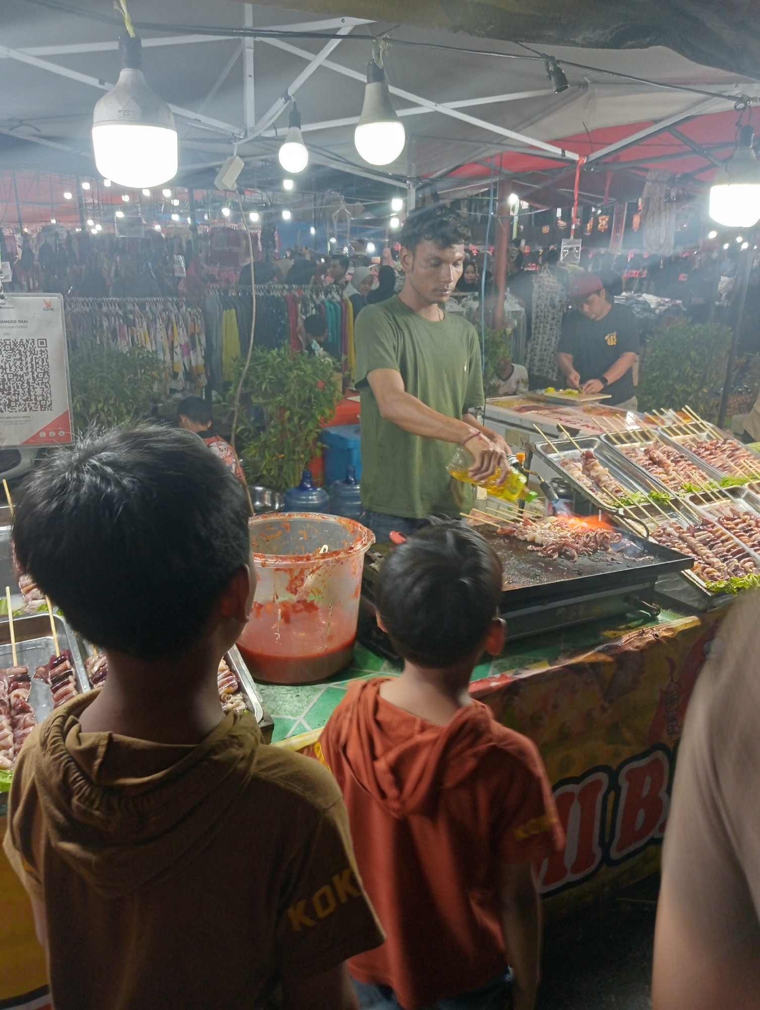 This is a nighttime, eye-level, medium shot capturing a scene at a food stall in Jambi, Indonesia. The focus is on a man in a green t-shirt grilling skewers of meat over a flat-top grill. He is pouring oil from a yellow bottle onto the grill, causing flames to erupt. In the foreground, two young boys are looking towards the grill, their backs to the camera. The boy on the left wears a brown hoodie with the word "KOK" visible on the sleeve. The boy on the right wears an orange hoodie. Behind the grill, other food items on skewers are displayed in trays. Various tents and lights suggest a busy night market or street food area. In the background, there are more stalls with clothing racks and people browsing. The environment appears to be an outdoor, urban setting with a humid, possibly smoky atmosphere.
