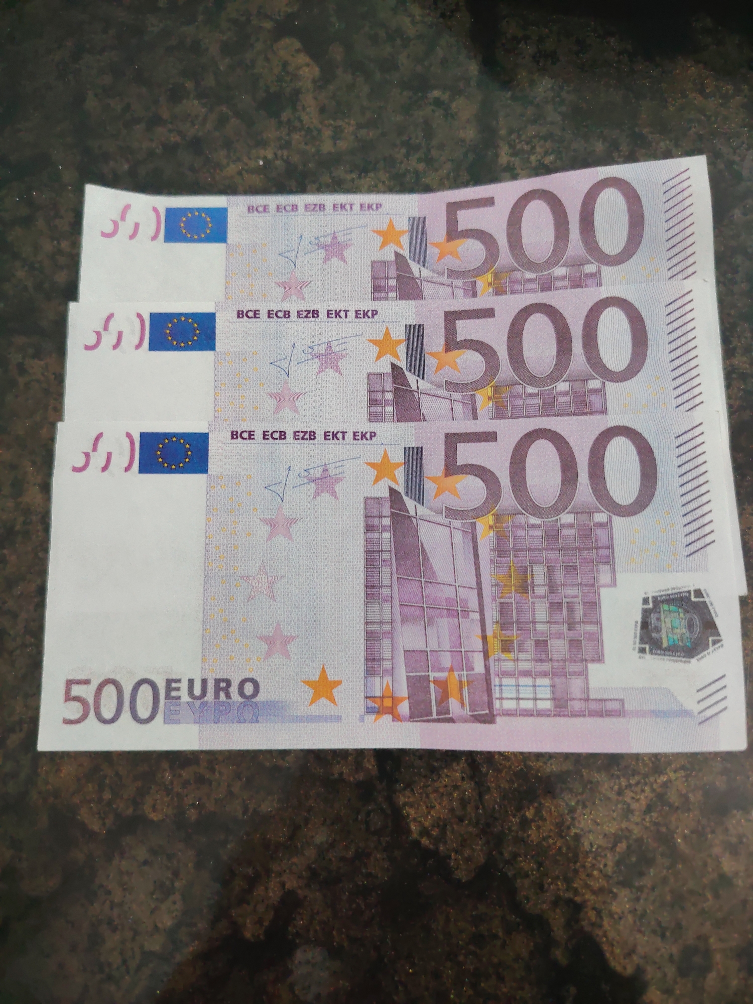 Here are three 500 Euro notes stacked on top of each other. The notes are predominantly purple and white in color. The value "500" is printed in large numerals on the right-hand side of each note. On the left side of each note is the word "EURO." The notes also feature a building facade. They are on top of a dark mottled surface.