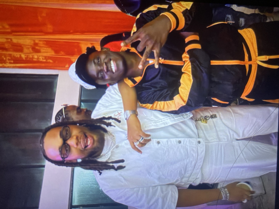 Two men smile broadly. The man on the bottom wears white pants, jacket, and sneakers. He has long dreadlocks, glasses, and multiple rings and bracelets. The other man wears a black and orange tracksuit, white hat, and silver watch. Both men are dark-skinned. They appear to be posing for a picture. The background is mostly white and orange.