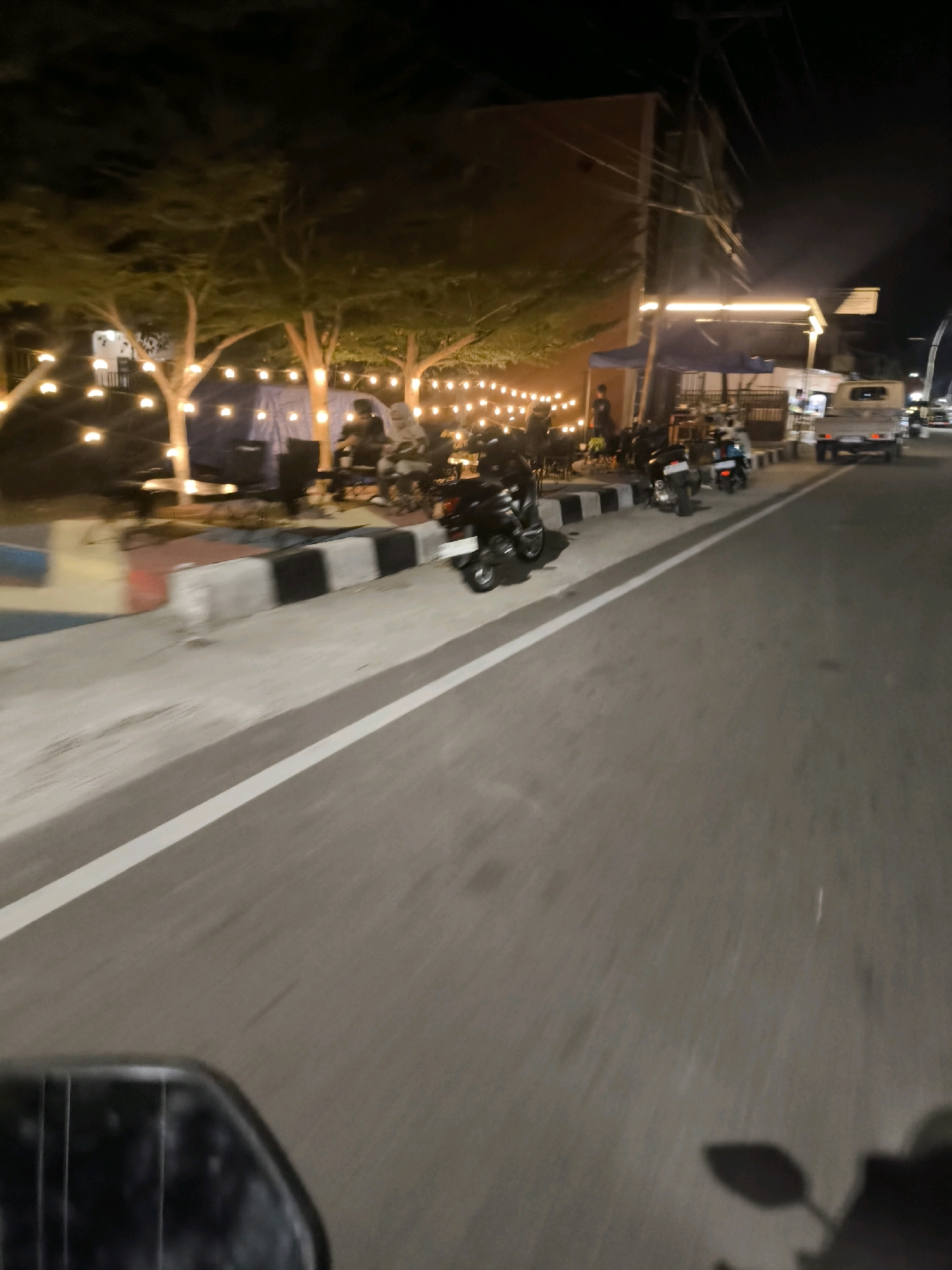 The image shows a street scene at night in Kwandang, Indonesia. Several motorcycles are parked along a sidewalk decorated with strings of lights. A few people are seated at tables in an outdoor seating area, illuminated by the lights. A small truck is visible on the road. The overall impression is of a small gathering or cafe.