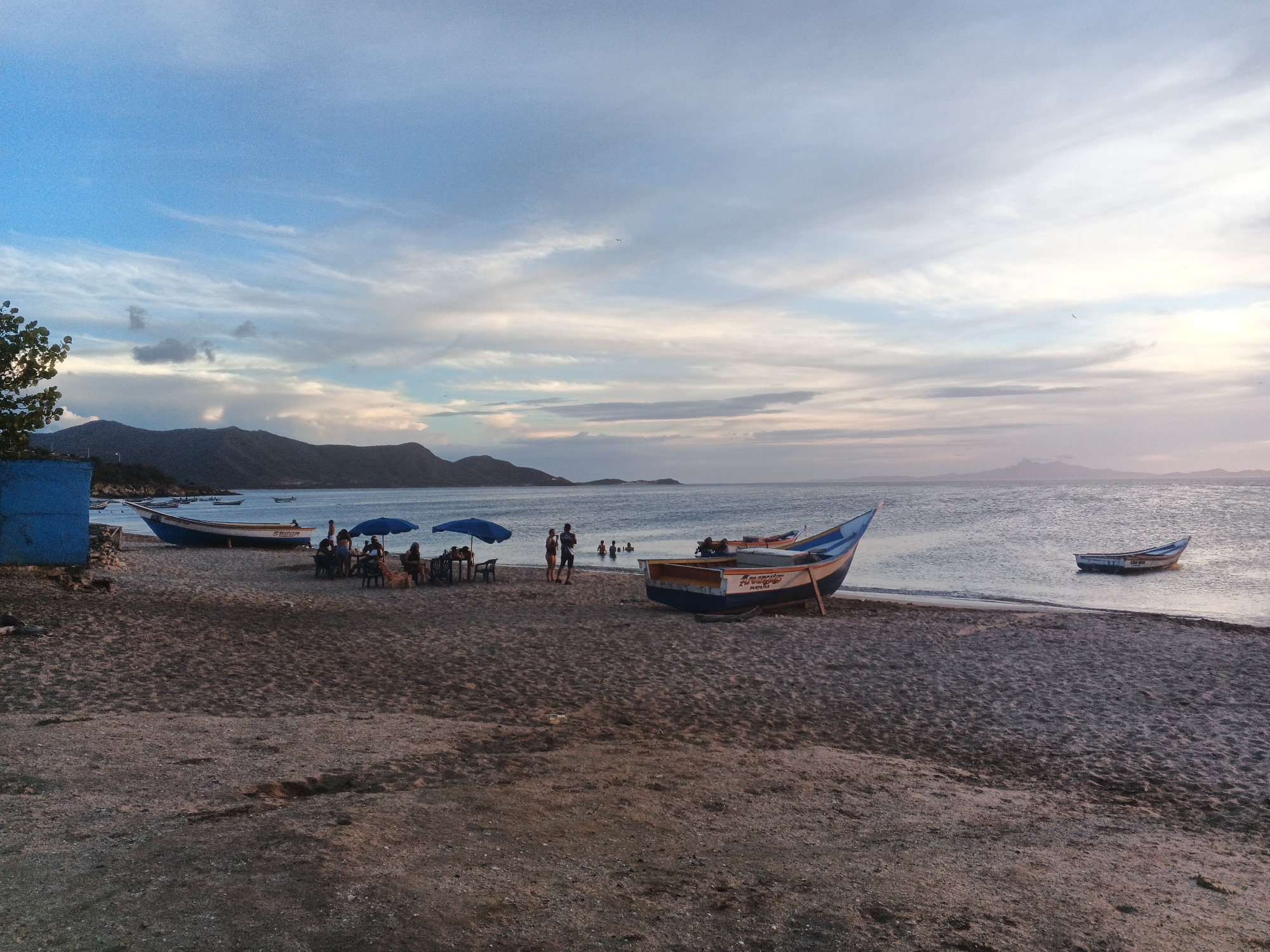 The image depicts a tranquil beach scene at dusk. The sky is a gradient of blues and whites, with wispy clouds suggesting a gentle breeze. In the distance, hazy mountains form a silhouette against the horizon. The foreground is dominated by a sandy beach, with ripples of sand indicating the tide's ebb and flow.
Several boats are visible: two are beached on the sand, one larger with blue and white detailing, and a smaller one further out in the water. Another long, narrow boat is also beached, possibly used for fishing. A cluster of people are gathered near the beached boats, some seated at tables under blue umbrellas, suggesting a small dining or resting area. Others are standing or wading in the shallow water. A blue structure, possibly a building or shed, is located on the far left of the image. The overall atmosphere is peaceful and serene, typical of a quiet evening by the sea.