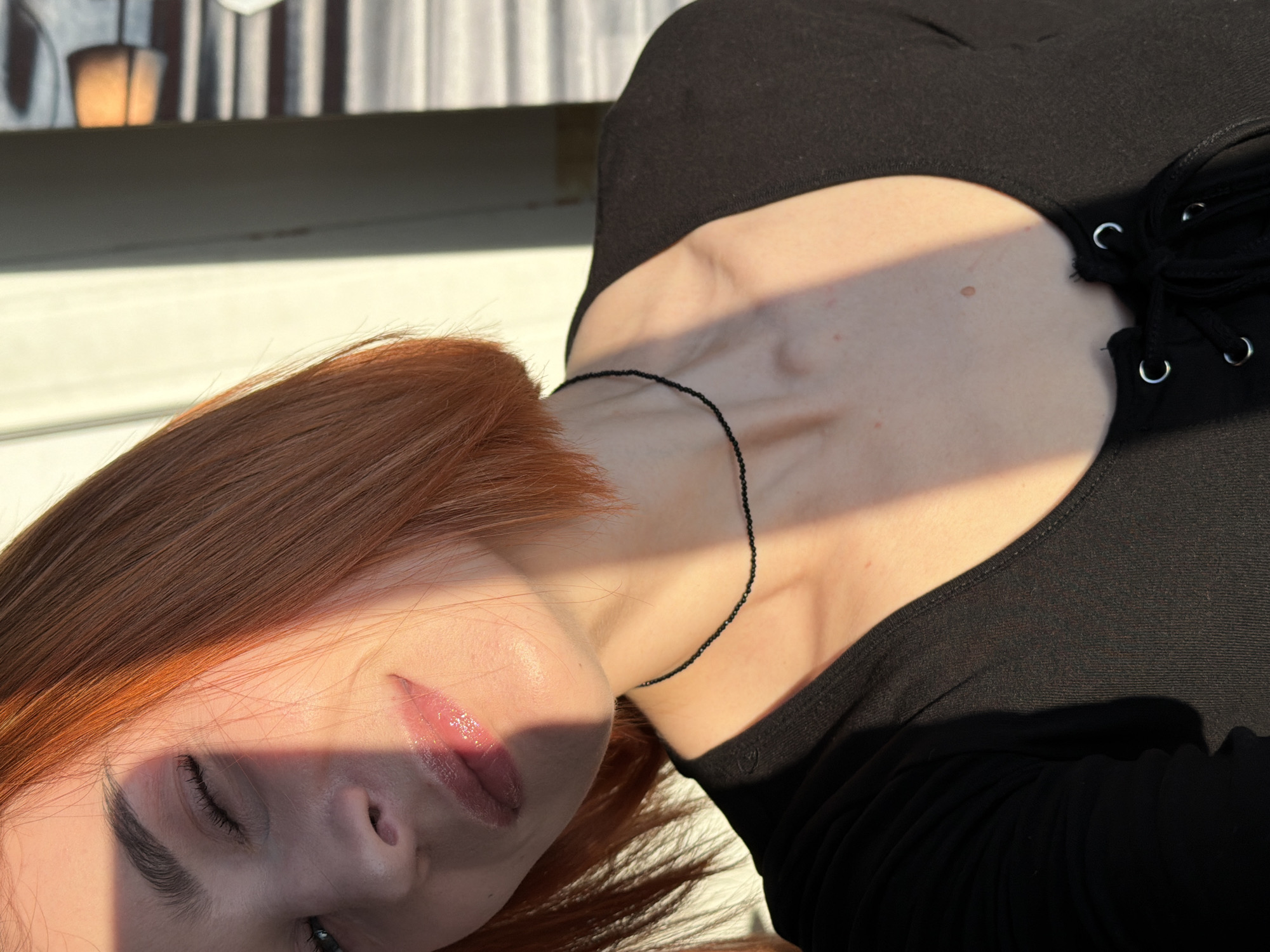 A close-up, slightly angled photograph depicts a person with red hair, closed eyes, and glossy lips. The person is wearing a black top with a cut-out neckline that is laced. A thin, black necklace is visible around the person's neck. Sunlight casts a strong shadow across the person's face and chest. The background appears to be a blurred outdoor setting, possibly a balcony. No information about the location in the city of Chaikovsky, Russia, is detectable from the image.