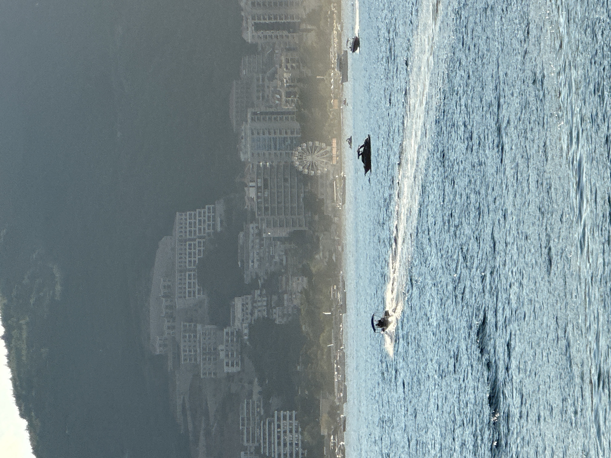 In the foreground, the bright blue water of the Adriatic Sea is rippled by the movement of several motorboats. One boat, with a dark canopy, speeds across the frame, leaving a white wake. Further in the distance, other boats are visible, some anchored and some in motion. The middle ground shows a coastline with a Ferris wheel and numerous modern buildings, including apartment complexes and hotels, built on a hillside. The buildings are a mix of white and light-colored facades, with many windows visible. The background is dominated by a large, tree-covered mountain, shrouded in a slight haze. The overall scene depicts a sunny day at a coastal city.