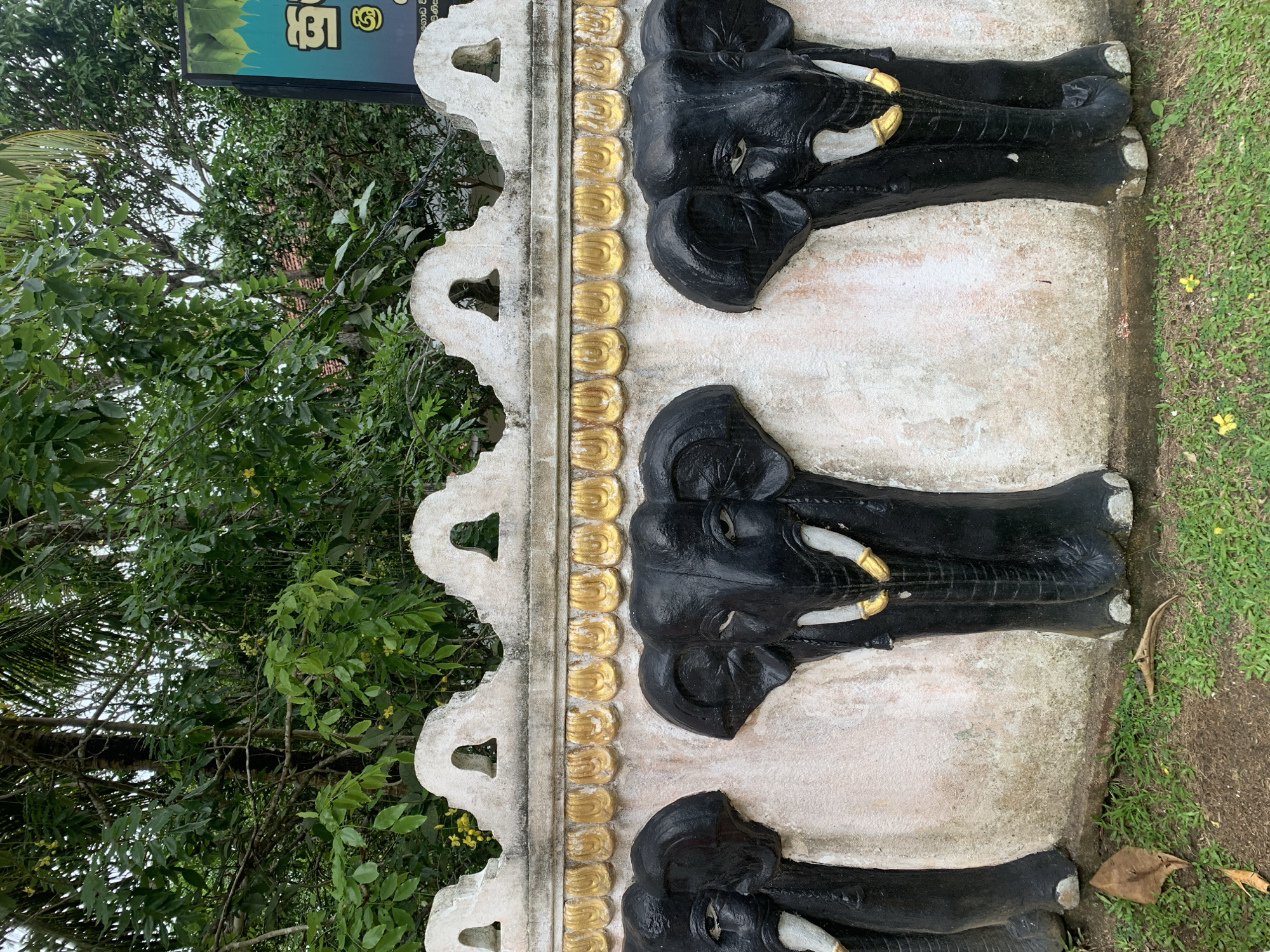 The image shows a decorative wall with three black elephant head sculptures protruding from it, located in Kabalana, Sri Lanka. The wall is made of a light-colored material and features a decorative gold trim above the elephants. The elephant sculptures are detailed, with visible tusks painted white and gold accents. Behind the wall, lush green trees and foliage are visible, suggesting an outdoor setting. A sign with text in a non-English script is partially visible in the upper right corner of the image. The ground in front of the wall is covered with grass and a few fallen leaves.