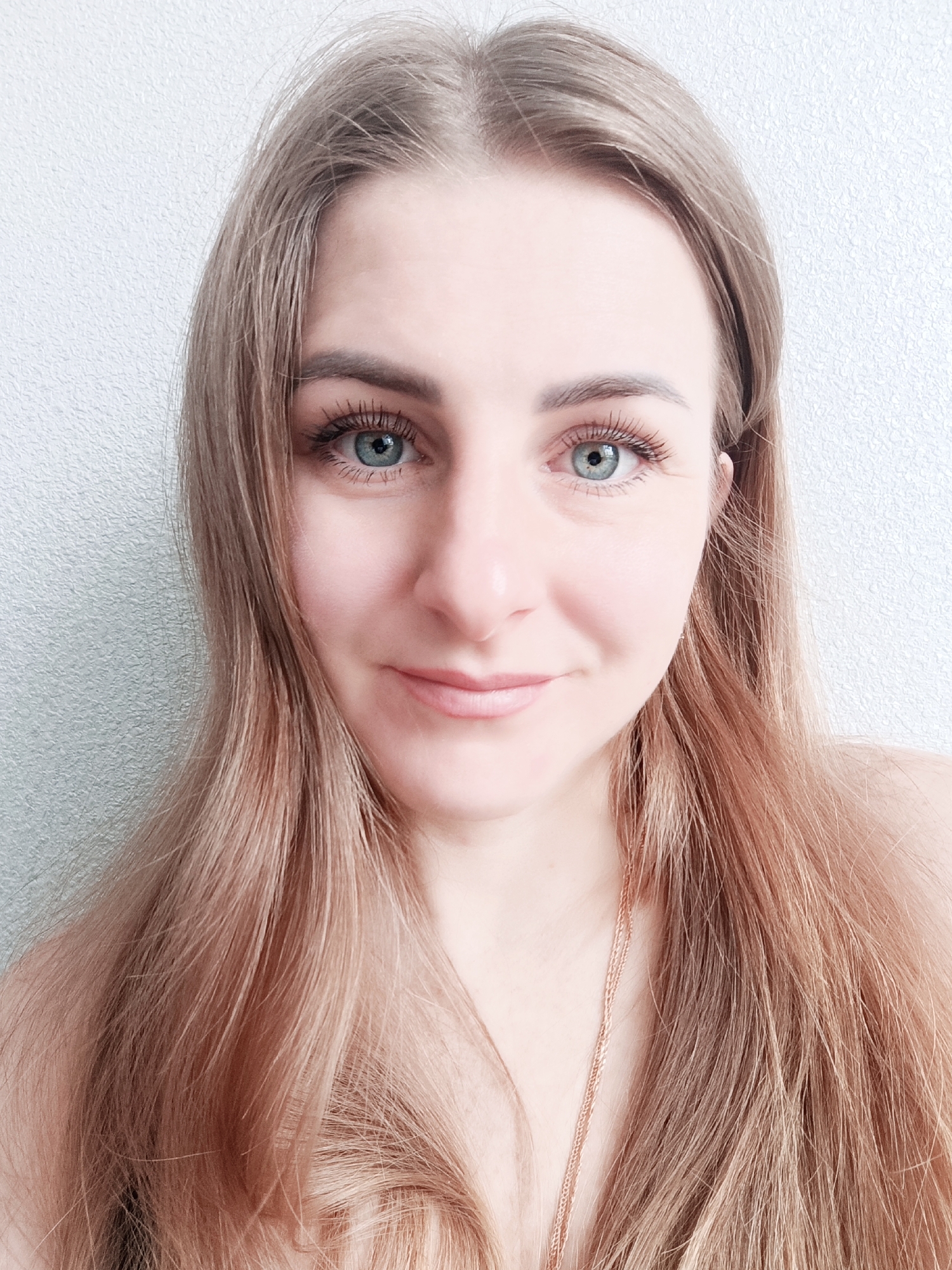 A close-up portrait features a young woman with long, light brown hair styled straight, falling over her shoulders. She looks directly at the camera with a gentle, pleasant expression and a subtle smile. Her eyes are a striking blue-green, enhanced with visible mascara on her eyelashes. Her skin is fair, and her lips appear to have a natural or lightly colored gloss. A thin, delicate gold or rose gold chain necklace is visible around her neck and extends down her chest, suggesting she is wearing a low-cut top or is bare-shouldered. The background is a plain, light-colored wall with a subtle textured finish, possibly stucco or a similar uneven surface, providing a simple and consistent backdrop. The lighting is soft and even, highlighting her features without harsh shadows, indicative of an indoor setting. There is no visible text in the image. The time of day and specific location are not discernible.