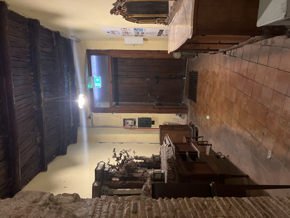 The image shows a hallway interior, located in Naples, Italy, at coordinates 40.852, 14.257. The hallway is dimly lit with one lightbulb hanging from the ceiling. The ceiling is made of exposed wooden beams. The walls are painted yellow. The floor is made of red tile. There is a wooden door at the end of the hallway. The door has a metal grate in front of it. There are two pieces of furniture on each side of the hallway. One piece is a dark wooden cabinet with a marble countertop, and the other is a smaller, wooden table with drawers. There are a few items on the table, such as a small stool and a plant. There is a white box sitting on the floor near the cabinet on the right. The hallway appears to be empty and there are no people present.