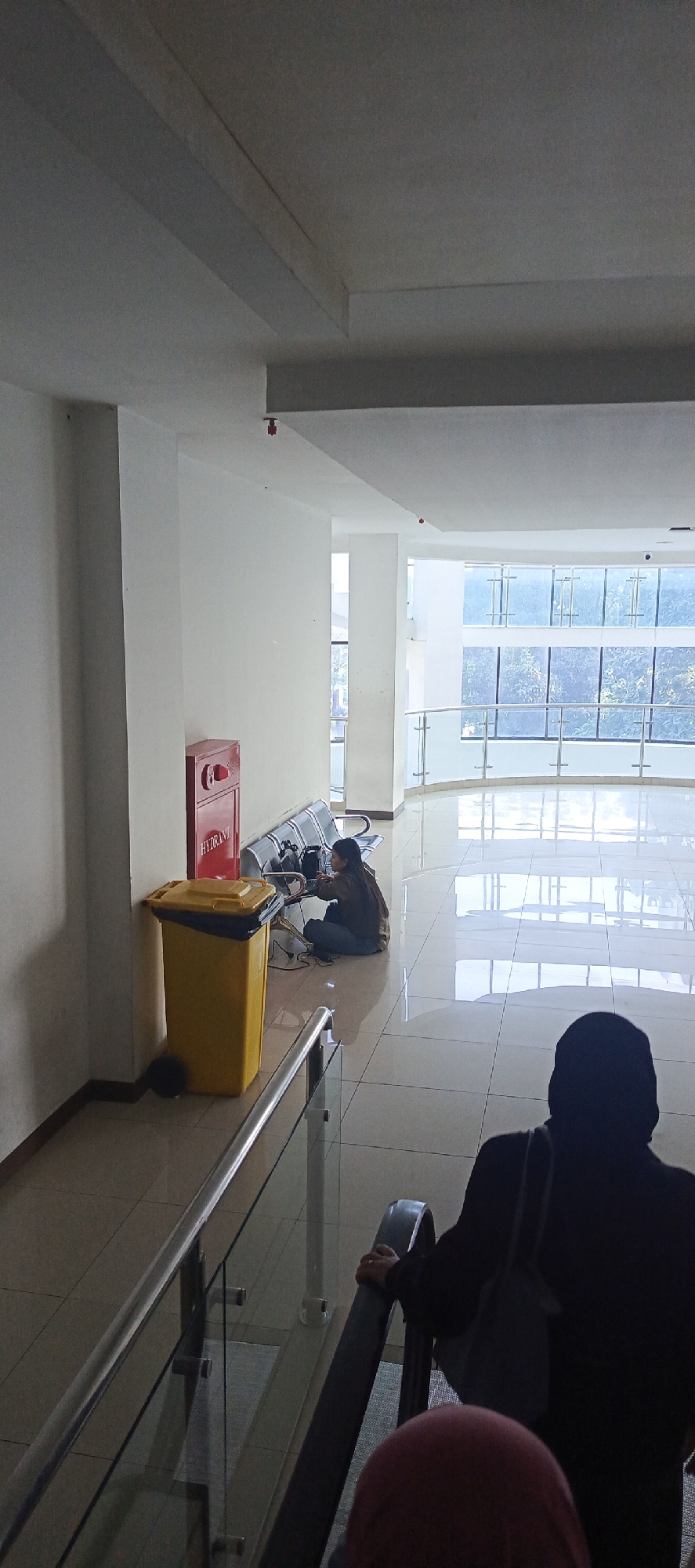 The image depicts an indoor scene, likely a public building lobby or waiting area in Jayamukti, Indonesia. The environment is characterized by large windows, a reflective tiled floor, and white walls. In the foreground, two individuals are partially visible, one on an escalator and another seated on the escalator's floor. In the mid-ground, a person is seated on the floor next to a yellow trash can and a red fire hydrant. The person appears to be working on a laptop, with cables extending from it. There are also several dark-colored chairs lined up against the wall. The overall impression is of a functional, modern indoor space.
