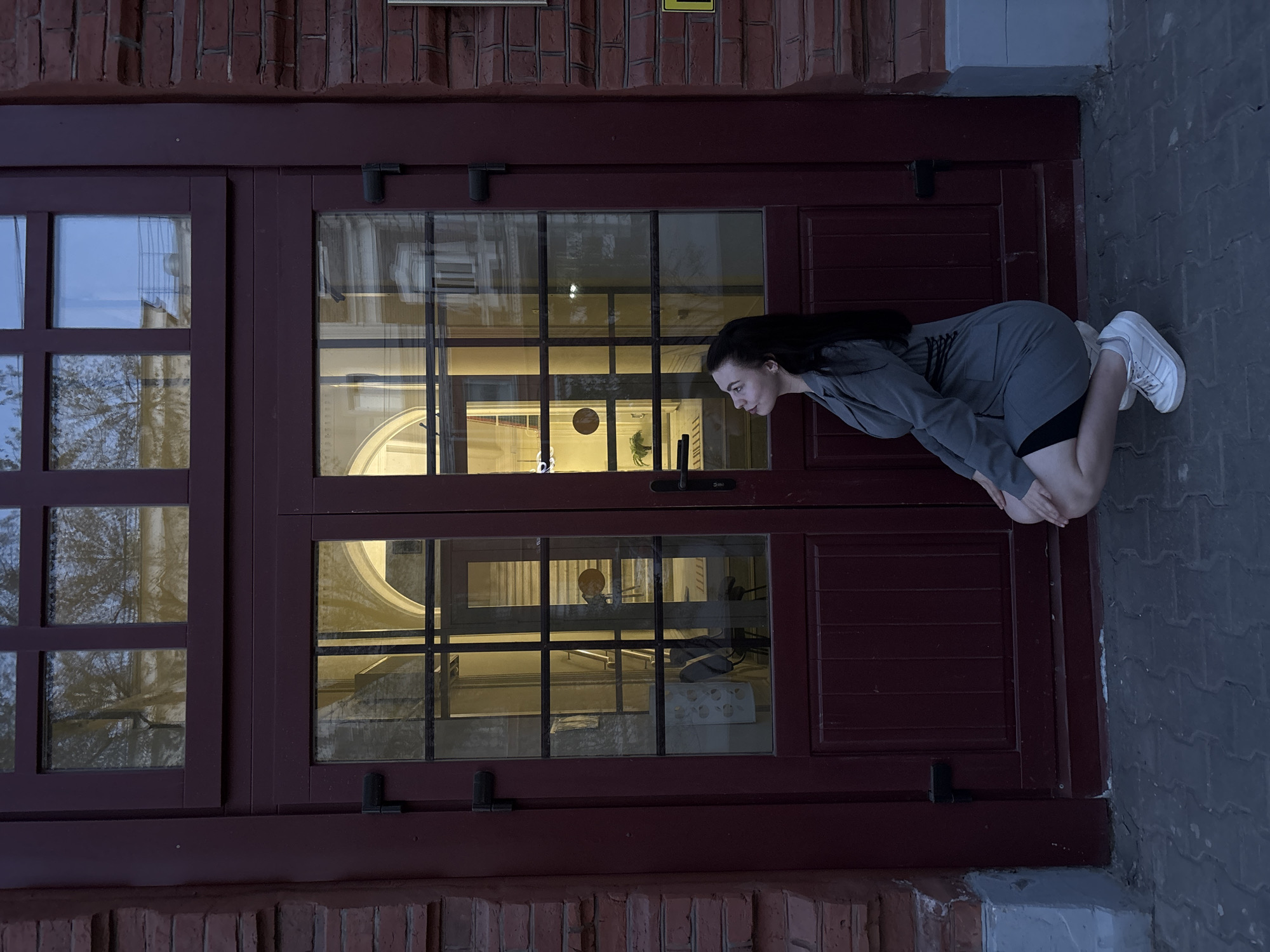 A woman with long, dark hair, wearing a gray blazer, black shorts, and white sneakers, is crouched down and appears to be peering into a large, dark maroon-colored window. The window is part of a building constructed of brick and features a grid pattern. The reflection inside the window suggests an interior lit with warm light. The scene is outside on a paved surface made of gray bricks. Based on the reflections visible in the windows and the building's architectural style, this image appears to be taken in Perm, Russia.