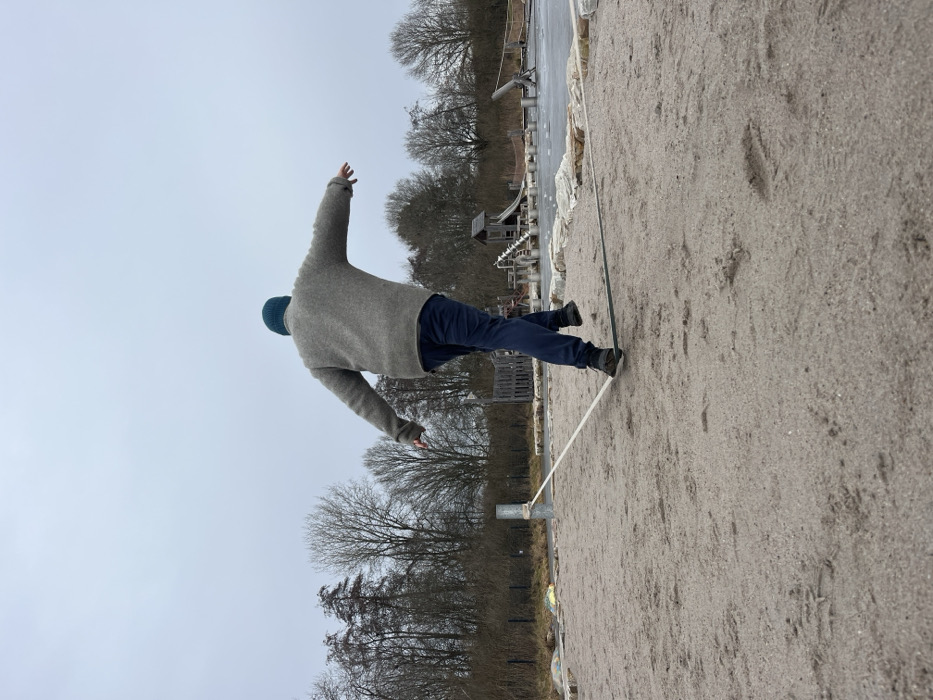 An individual is captured mid-action on a slackline in an outdoor recreational area in Nohfelden, Germany. The subject, viewed from behind, wears a teal knit beanie, a grey textured sweater, dark blue pants, and dark athletic shoes, with arms extended horizontally for balance. Their left foot is positioned on a low-lying slackline, while the right foot is elevated above it, indicating a step. The slackline, composed of white and dark green sections, is anchored to a grey metal post on the left. The ground in the foreground and midground consists of light tan sand with visible footprints. In the background, a paved path, a wire fence, and numerous bare deciduous trees are visible. To the right, playground equipment including a wooden structure resembling a hut with a slide, along with various stone elements, and a dark, flat area, possibly an ice-covered pond or rink, complete the scene under an overcast sky.