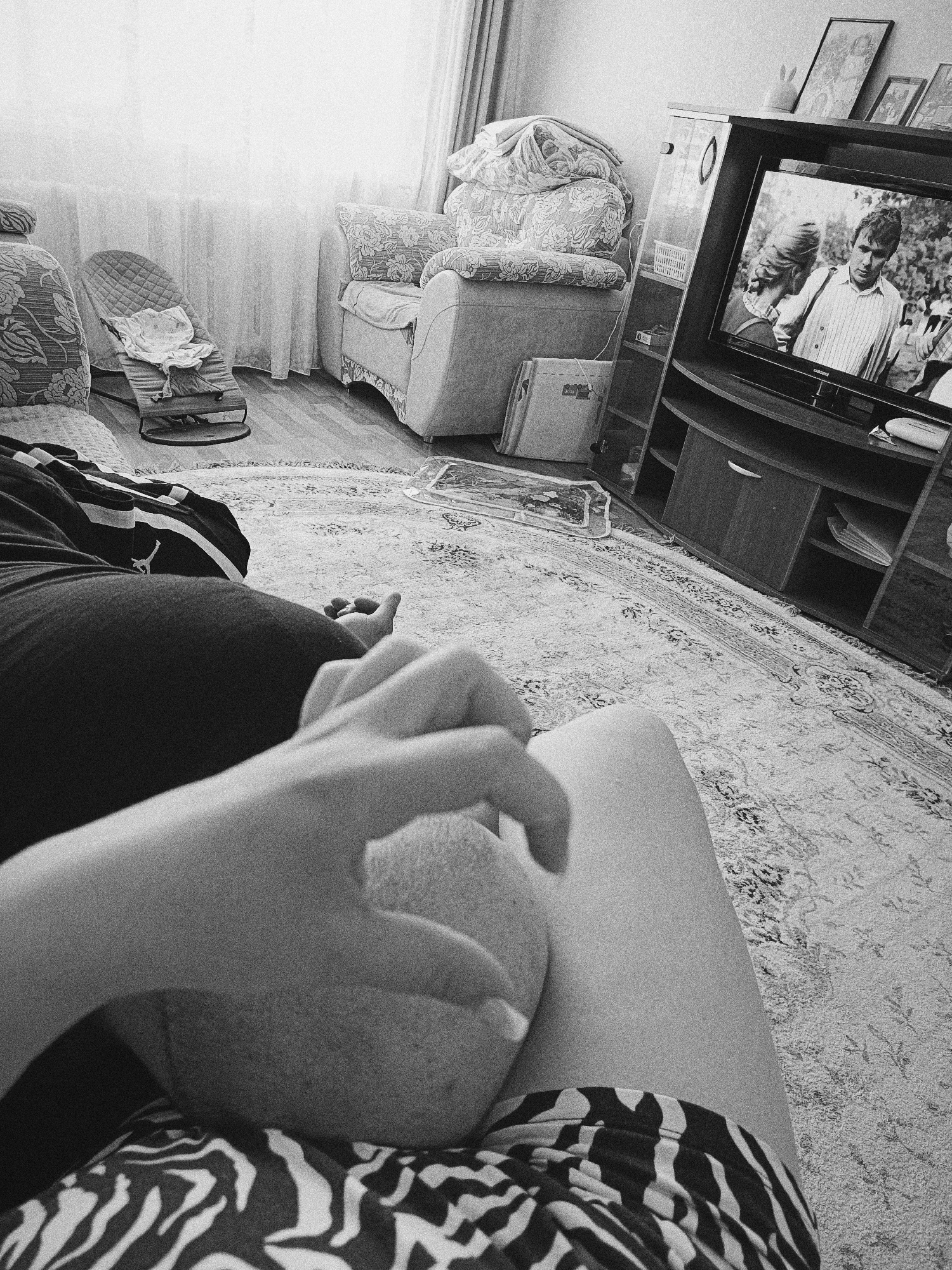 The photograph is taken indoors. A person is sitting on a couch, wearing a black shirt and shorts with a black and white zebra print. The person is holding an apple. In the background, there is a television set, a baby rocker, and a floral-patterned chair. The television is playing a black and white movie. The location is in a room with a patterned rug in what appears to be a home; the location details regarding Tobolsk, Russia are not evident in the image.