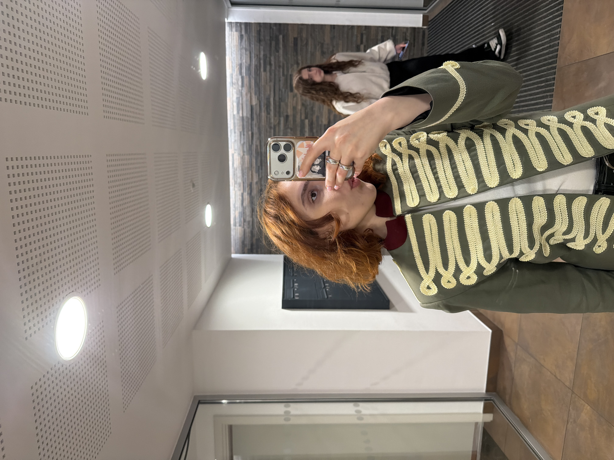 The image shows a young woman with reddish-brown, curly hair taking a selfie in a hallway. She is wearing a green jacket with elaborate gold braiding and a dark red collar. She has two silver rings on her left hand, which is holding a smartphone with a case featuring pictures of people. In the background, another young woman with long brown curly hair is visible, wearing a cream-colored top and black pants. The hallway has a tiled floor, white walls, a black wall-mounted unit, and a stone-patterned wall. The ceiling is white with a grid of small square perforations and recessed circular lights. The scene is indoors, likely in a residential building or a public corridor. There is no indication that this location is in Strasbourg, France.