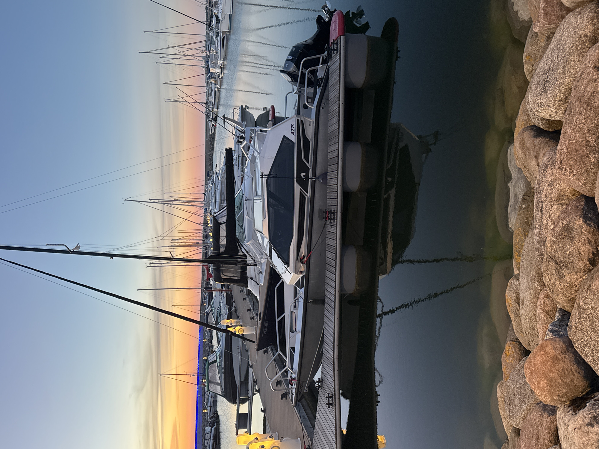The image shows a marina at dusk, with several boats docked. In the foreground, a modern motorboat with a silver hull and white cabin is moored to a wooden dock. The boat has a Suzuki outboard motor. Behind it, a darker motorboat with a green-tinted canopy is visible. Further back, numerous sailboats with tall masts are lined up in the marina, their rigging silhouetted against the sunset sky. The sky transitions from a warm orange and yellow near the horizon to a soft blue overhead. The water reflects the boats and the sky. In the very foreground, a line of large, rough-hewn rocks forms a barrier between the water and the viewer. The scene suggests a peaceful evening in a coastal city. This scene is not specifically identifiable as Tallinn, Estonia, but it depicts a common type of harbor found in many European coastal cities.