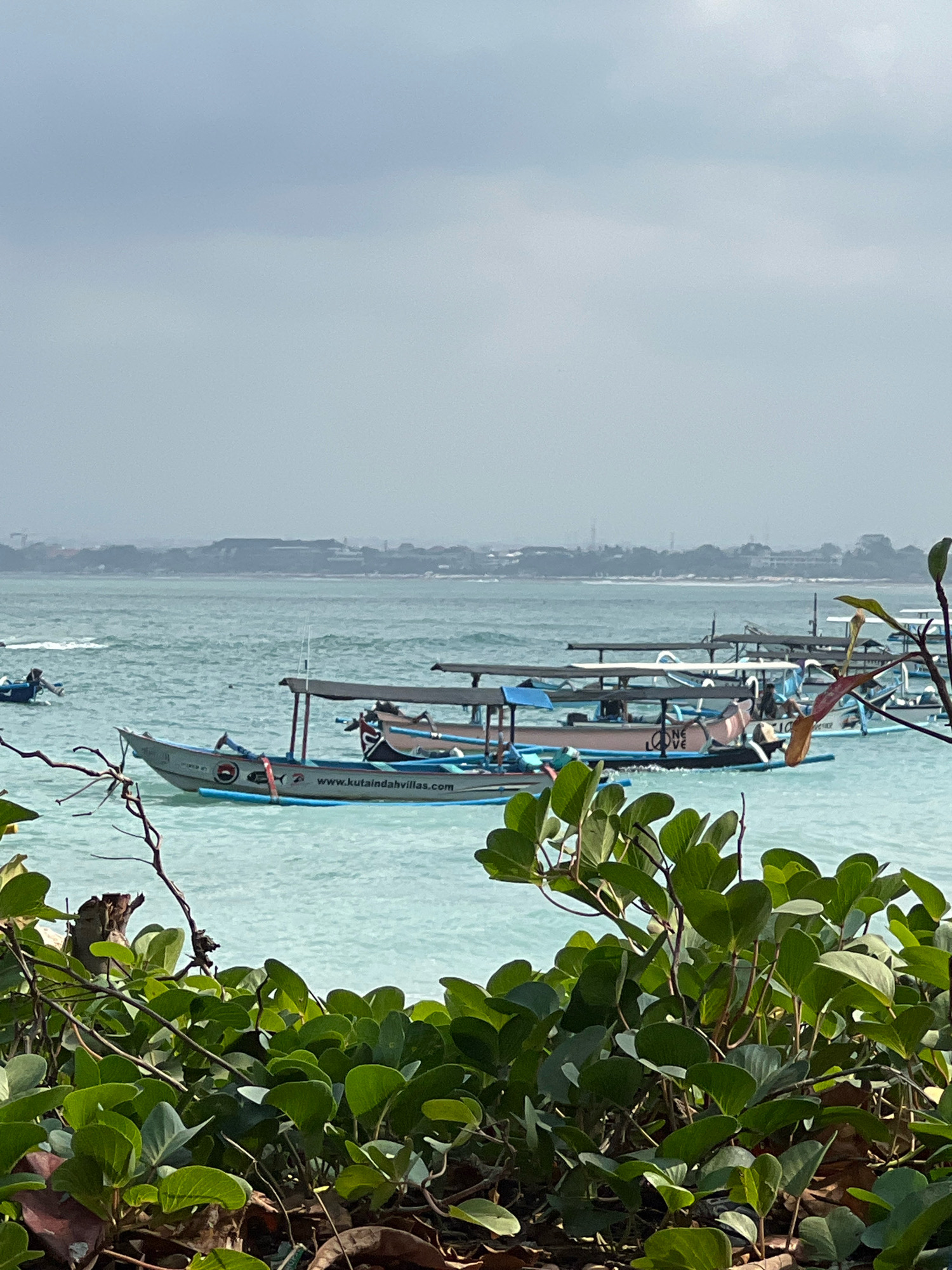 A serene, overcast day scene unfolds at Kuta Beach, Bali, Indonesia, with a focus on the tranquil waters and several traditional Balinese outrigger boats. The foreground is dominated by lush, green foliage, obscuring the sandy shore and suggesting a perspective from slightly elevated ground or a beachside planting.
In the middle ground, the shallow, turquoise water is dotted with several of these distinctive boats. The closest boat, a narrow, silver outrigger, prominently displays a website address: "www.kutaindahvilllas.com". It also features graphics of a fish and a circular logo. Another boat further back has the words "ONE LOVE" painted on its side, with additional passengers visible within its shaded interior. Other boats are partially visible, some with their characteristic outriggers extending outward. The water is calm, with gentle ripples reflecting the muted sky.
In the background, a hazy, indistinct coastline stretches across the horizon. Silhouettes of buildings and perhaps some industrial structures are barely discernible through the atmospheric haze, indicating the urban presence beyond the beach. The sky is a uniform pale blue-grey, suggesting overcast conditions or a light mist. The overall impression is one of peaceful coastal repose, characteristic of many areas in Kuta.