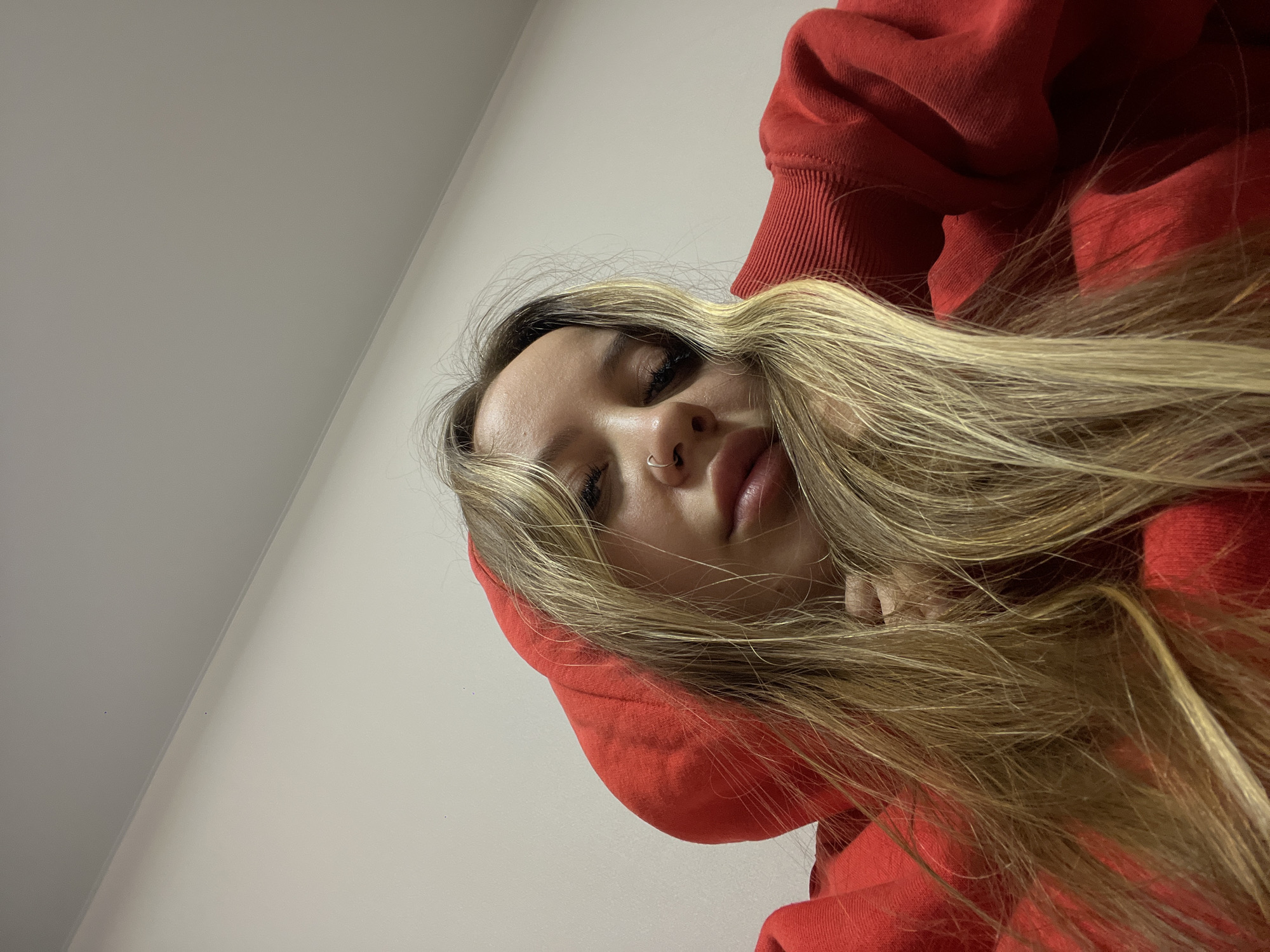 A young woman is captured in a close-up, tilted portrait, likely a selfie. She has long, wavy blonde hair with darker roots, which is spread out around her head and over her shoulders. She is wearing a vibrant red hooded sweatshirt, with the hood seemingly pulled up and framing her face. Her eyes are partially closed, giving her a relaxed or perhaps sleepy expression, and her lips are slightly parted. A small, silver hoop nose ring is visible in her septum. The background consists of a plain light-colored wall or ceiling, divided by a subtle diagonal line, indicating an indoor setting with an unusual camera angle looking down or up at the subject. There is no visible text. The image does not contain specific cues to indicate the provided location context of Krasnodar, Russia.
