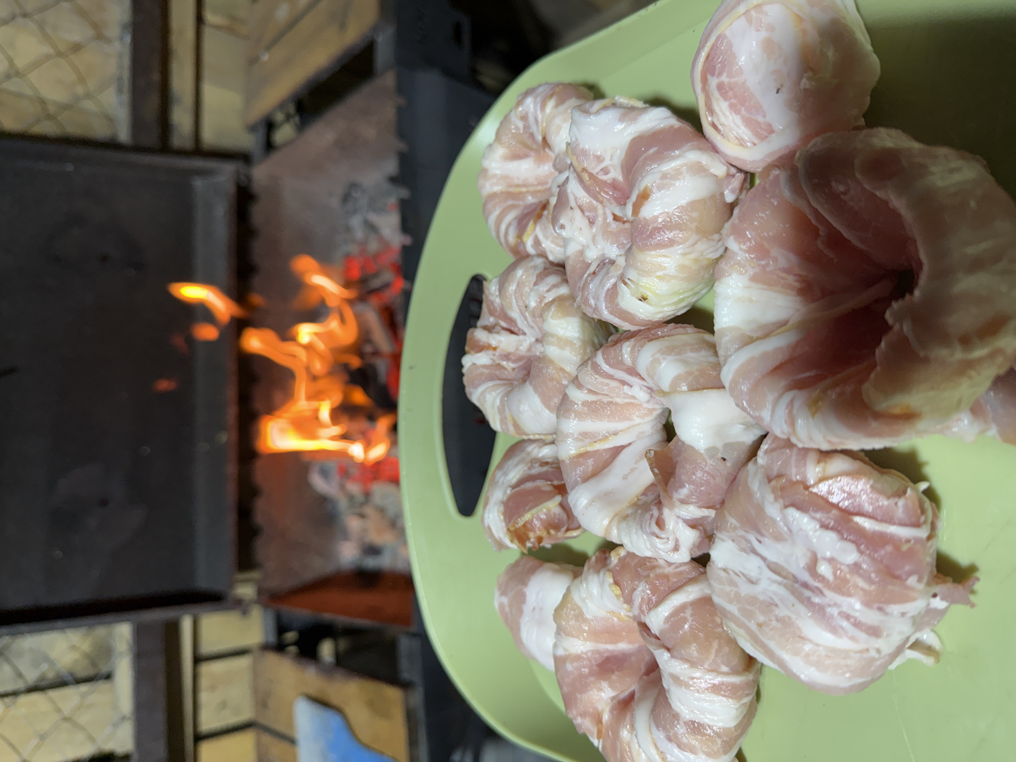 In an outdoor setting, likely a backyard or patio area, a green plastic tray holds several food items wrapped in bacon. These items appear to be circular with a hole in the center, resembling donuts or rings, and are arranged on the tray. In the background, a grill with glowing embers and active flames is visible, suggesting food preparation is underway. The overall environment suggests a casual outdoor cooking event. The location of Kamyshin, Russia is not discernible from the image.