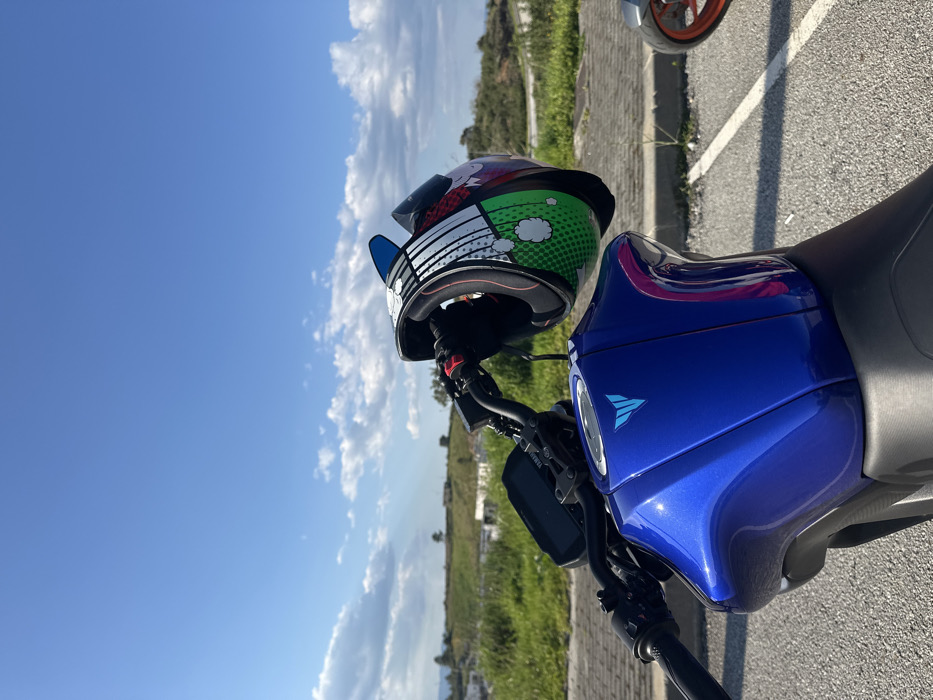 The scene depicts a metallic blue Yamaha motorcycle, identifiable by its fuel tank logo, parked outdoors in Mirandela, Portugal. A multi-colored full-face helmet with green, red, white, black, and blue comic-style graphics hangs from the motorcycle's black left handlebar. The motorcycle's digital instrument cluster and a portion of its black seat are visible. The ground consists of asphalt pavement, with a white parking line visible on the right. In the midground, a strip of green grass and weeds borders the paved area, leading to a landscape of dry grass, scattered bushes, and distant light-colored structures on a hillside. The sky is bright blue with scattered white cumulus clouds. A portion of a second motorcycle's front wheel, featuring an orange rim, is visible in the far right background.