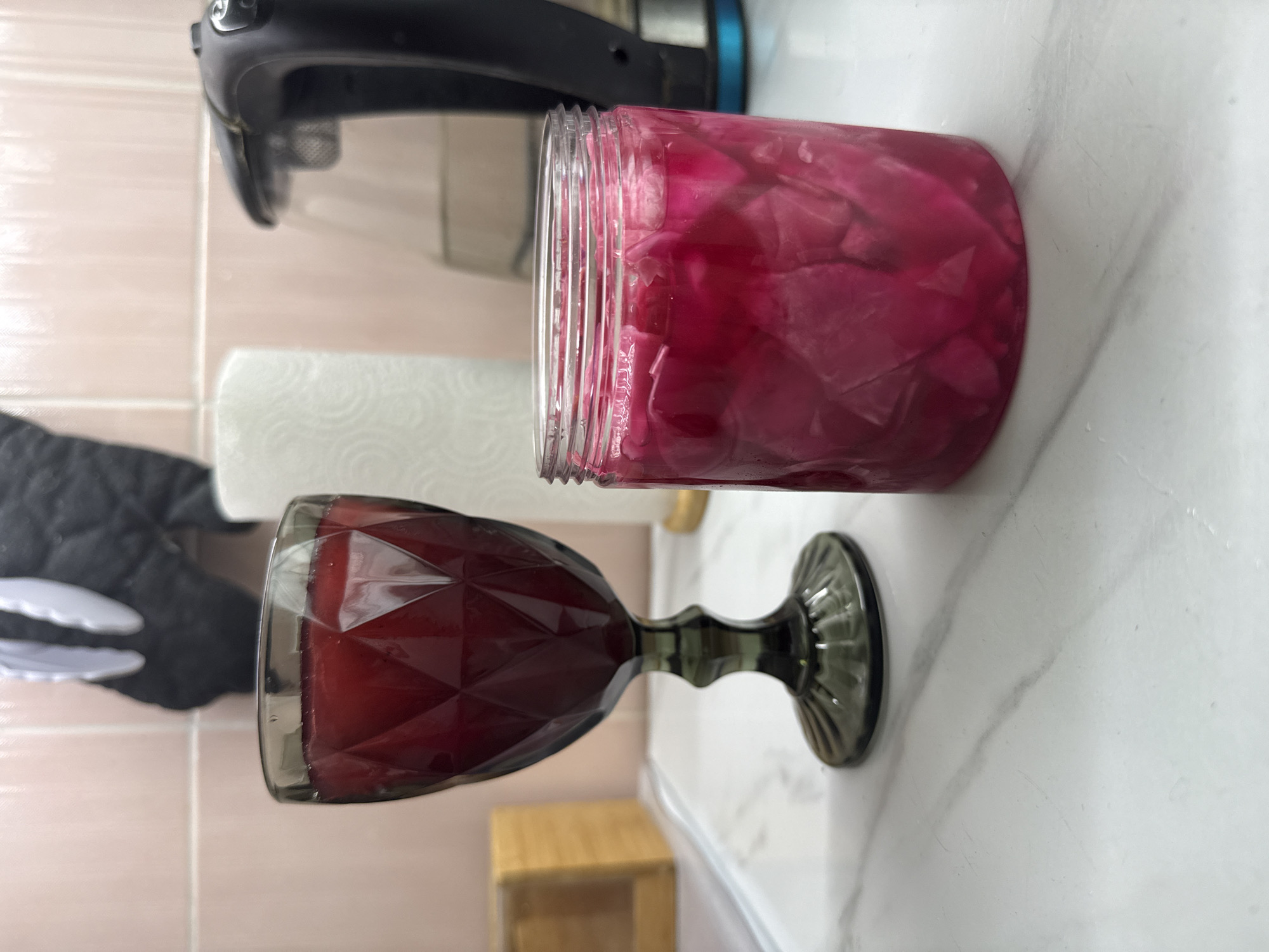 A horizontally oriented close-up shot depicts a kitchen countertop scene with various items. The countertop is white marble with grey veining.
A clear plastic jar filled with bright pink or red pickled cabbage sits near the center-right of the frame. Next to it, a dark red faceted wine glass rests on its side. A roll of paper towels is partially visible behind the jar, and a black and white oven mitt is on the left side of the frame. A black handheld vacuum cleaner is positioned in the upper left corner, mostly out of focus.
The image does not provide information to determine the specific location within Khimki, Russia.