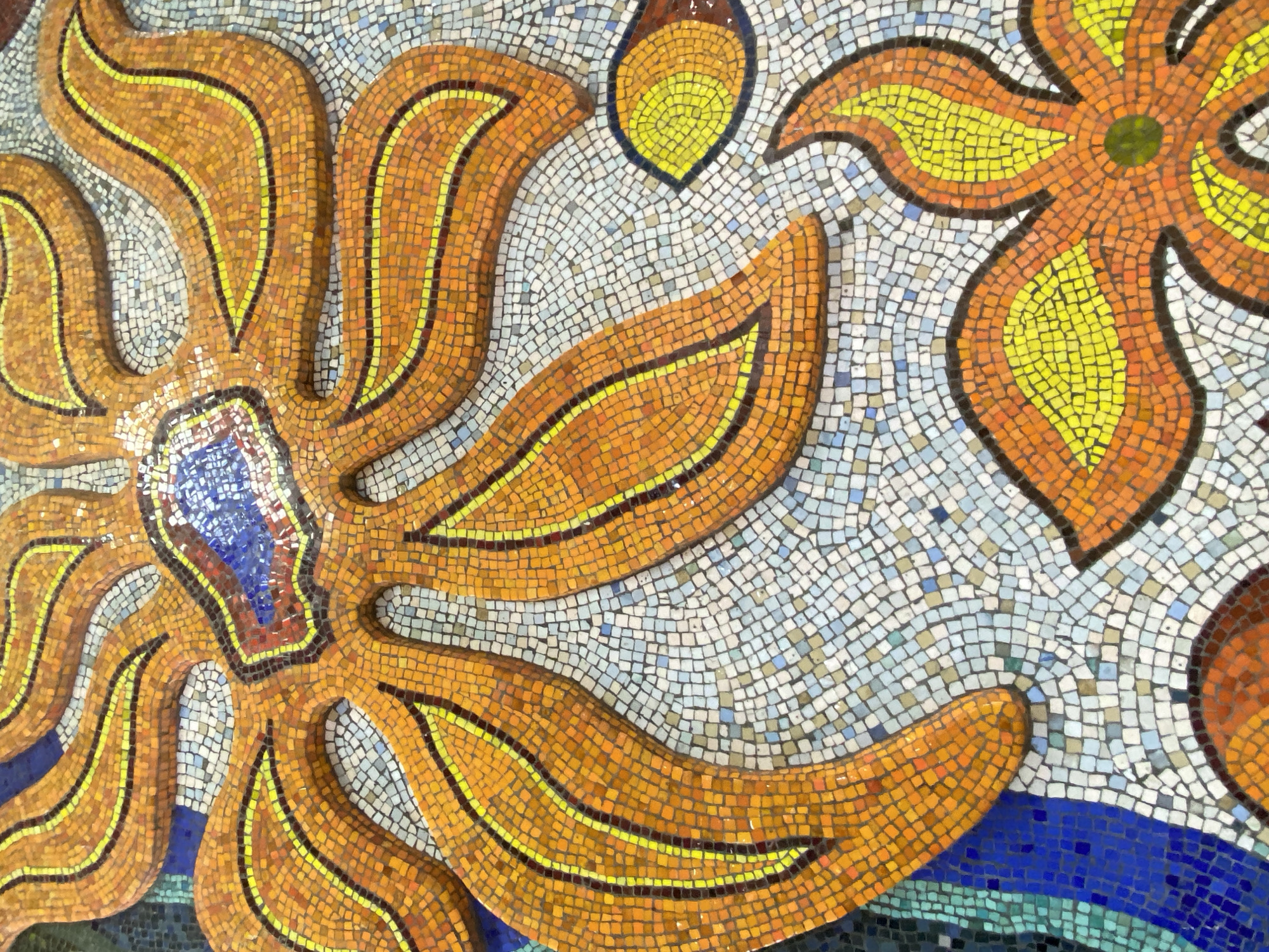 The image shows a close-up of a mosaic in Sochi, Russia. The mosaic features two stylized floral designs. The larger one, on the left, has long, curving orange petals with a yellow stripe running along each. The center of this flower has blue, red, and brown mosaic tiles. The second flower, on the right, has shorter, broader petals that are yellow with orange edges. The center of this flower is a green circle. The background around the flowers consists of small, square mosaic tiles in various shades of white, blue, and gray. Below the larger flower, there is a section of dark blue and turquoise mosaic tiles, suggesting water or another dark element. The mosaic appears to be part of a larger artwork, as the second flower is partially cut off. The composition is visually dense with small mosaic tiles.