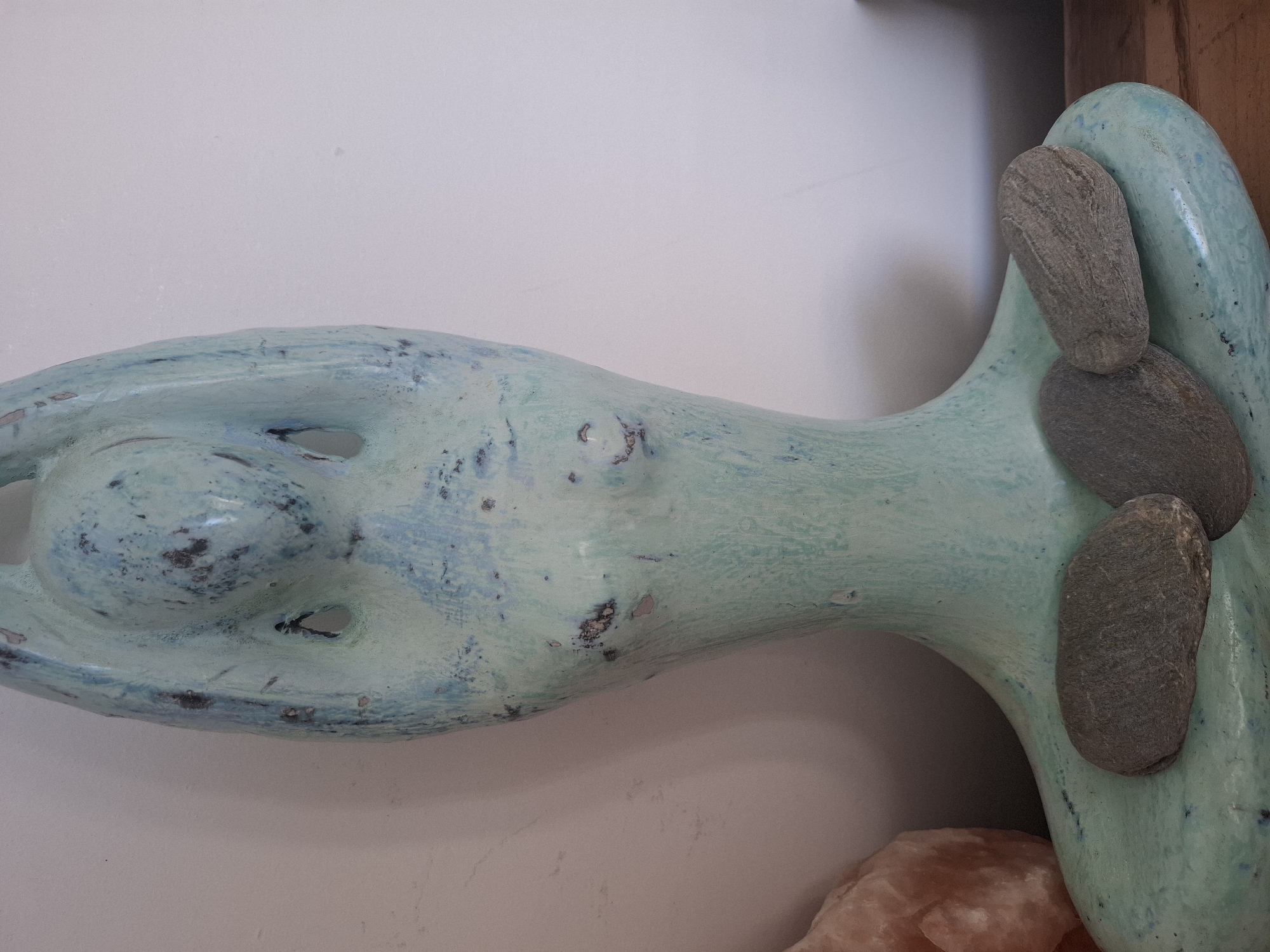 This image shows a close-up view of a light blue, ceramic statue with dark speckles, resembling a stylized human form in a seated position. The statue has two openings where the arms are bent upwards and hands are placed on the head. Three flat, grey stones are resting on the base of the statue. To the left of the statue, a large, irregular-shaped pinkish object, possibly a crystal or rock, is partially visible. The background is a plain, off-white wall. The scene is indoors, likely on a wooden surface. There is no discernible connection to the city of Montreuil, France, in the provided image.